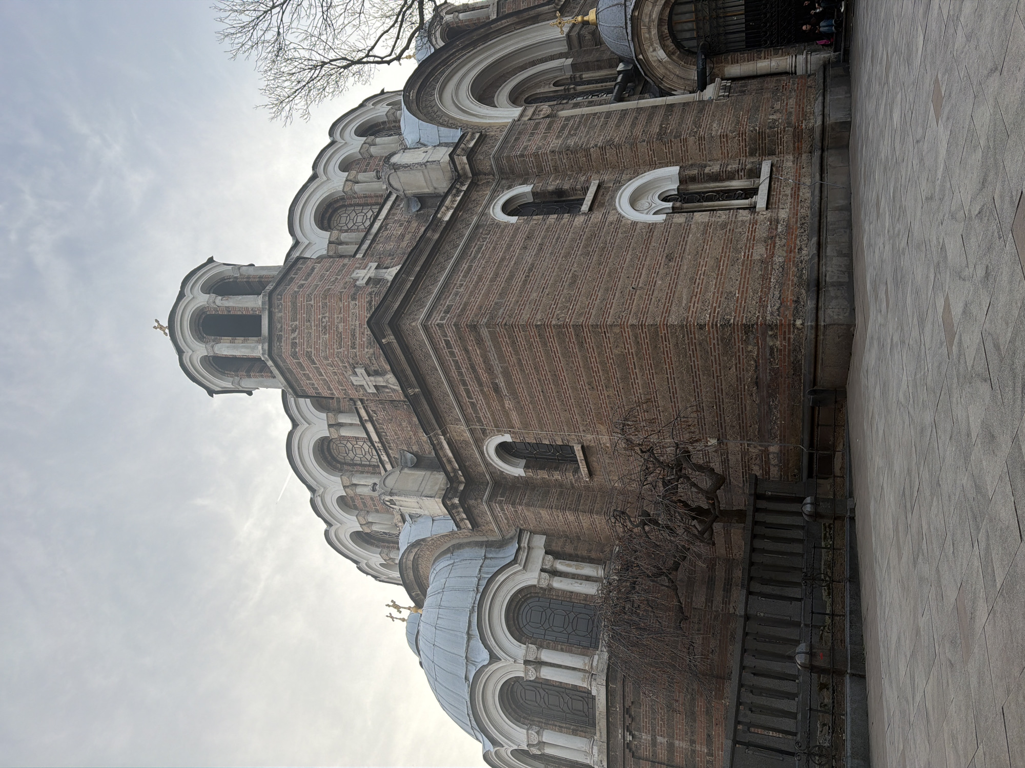The image shows a low-angle view of St. Alexander Nevsky Cathedral, a Bulgarian Orthodox cathedral in Sofia, Bulgaria. The cathedral features brickwork, arched windows, domes, and crosses on top. A bare tree is present on the right side of the frame. The sky is overcast with muted colors. The ground is paved. People can be seen near the cathedral's entrance.