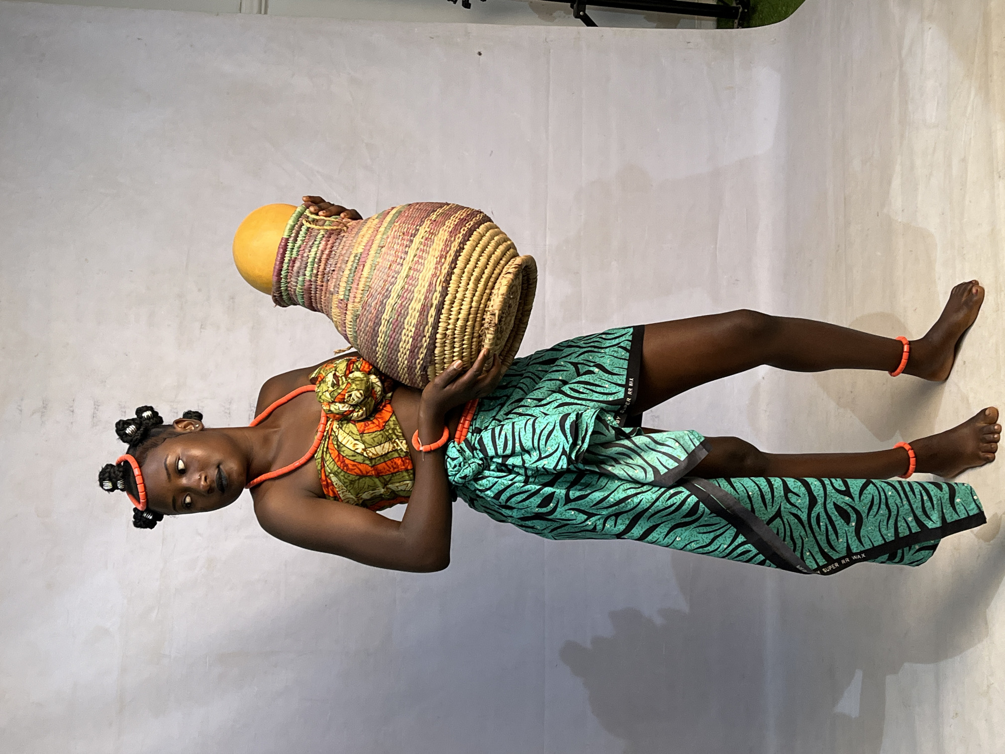 A full-body, medium shot shows a young Black woman with dark skin and braided hair styled in multiple buns, wearing bright orange beaded jewelry around her neck, wrists, and ankles. She is dressed in a vibrantly patterned green, orange, and yellow top and a matching patterned teal and black wrap skirt. Her left arm is bent as she holds a large, intricately woven straw basket containing a bright yellow object, possibly a fruit or gourd. The woman stands barefoot on a light-colored floor against a white, seamless backdrop. Shadows are visible on the left side of the image, and a subtle green element is present at the bottom right edge, suggesting an outdoor or studio setting with some controlled lighting. There are no discernible features that place the location specifically in Barakin Akawo, Nigeria.