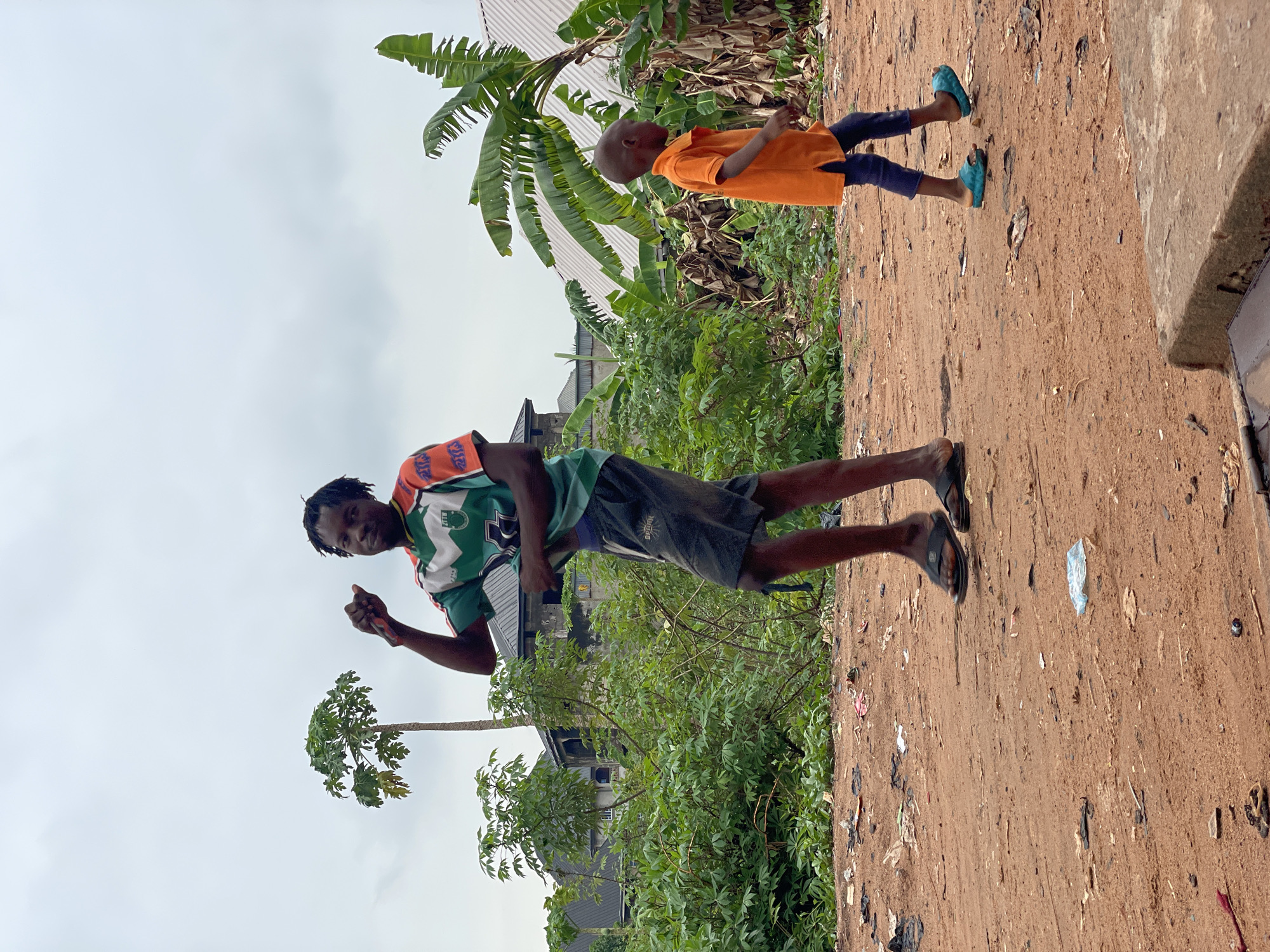 Two individuals are present in the image. An adult male, dressed in a patterned short-sleeve shirt and dark shorts, stands in the foreground. He is holding an object in his hand and is gesturing towards the camera. Behind him, a child is walking along a dirt path or road. The child is wearing an orange shirt, dark pants, and blue footwear. The surrounding environment features dense vegetation, including banana trees and other greenery. A building is partially visible behind the vegetation. The ground is dirt with visible debris and trash scattered about. The sky is overcast. The location appears to be a roadside in Onitsha, Nigeria.