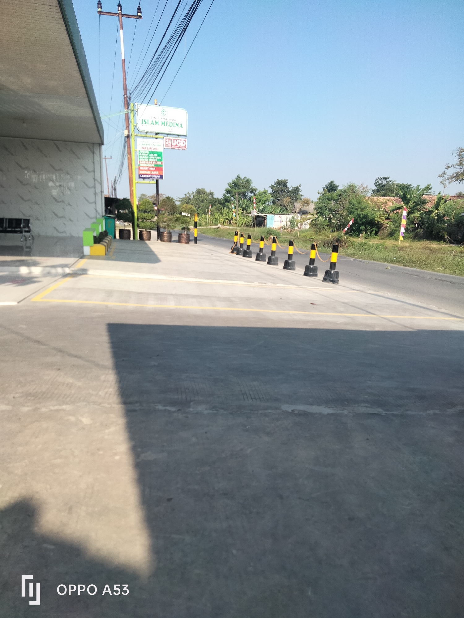 The image shows a street scene in Karawang, Indonesia, with a clinic sign visible. The sign reads "KLINIK UTAMA ISLAM MEDINA" and also indicates "24 UGD" and "MELAYANI." Several traffic cones are lined up along the side of the road, and a building with a covered walkway is on the left. The environment appears to be a somewhat rural or suburban area, with trees and vegetation visible in the background. The sky is clear and blue, indicating a sunny day. There are no people clearly visible in the image. The overall impression is one of a functional but modest facility on a roadside.