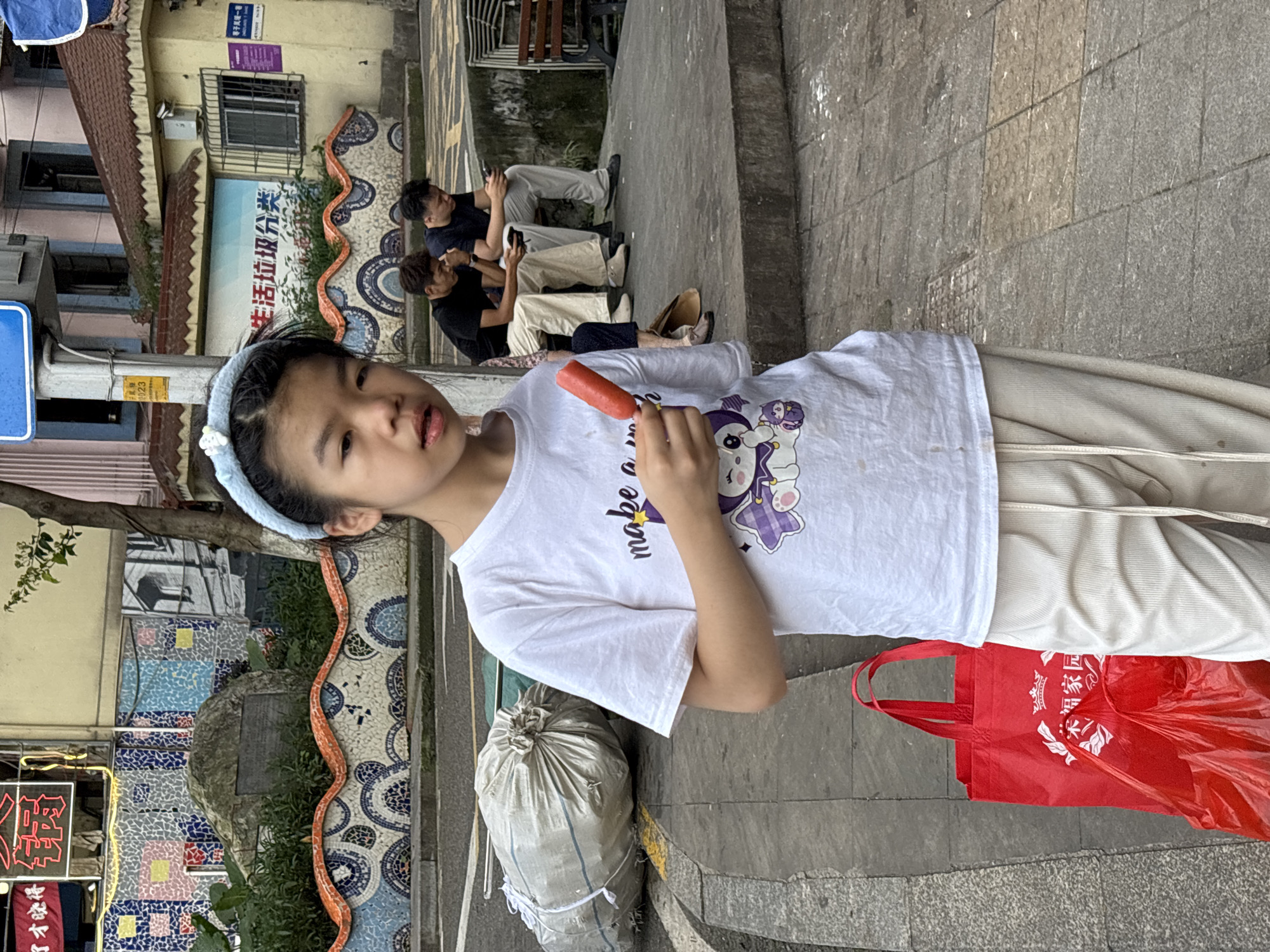 In the Yuzhong District of China, a young girl with a light blue headband and a white t-shirt stands on a tiled sidewalk, holding a red popsicle. Her t-shirt features a cartoon image of a cat-like character. She is wearing light-colored, loose-fitting pants. In the background, two men are seated on a stone ledge, both looking at their phones. Behind them, a building with a dark roof and yellow walls is visible, adorned with a colorful mosaic wall featuring blue, white, and orange patterns. A large, white, bound sack lies on the street to the left of the girl, and a bright red reusable shopping bag is placed on the sidewalk near her feet. A tree grows to the right of the mosaic wall.
