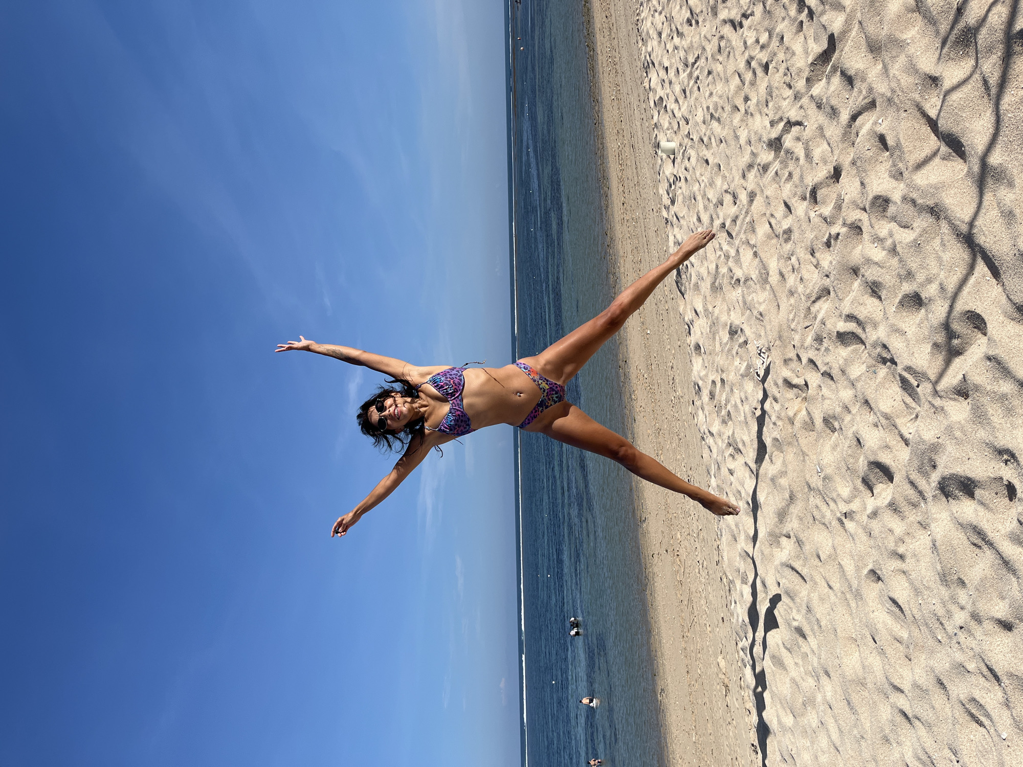 A young woman in a bikini lies on a beach in Kuta Selatan, Indonesia, with arms and legs outstretched, against a backdrop of blue sky and ocean. The sand is light-colored, and the ocean is calm. There are a few people in the water. The photo is oriented sideways.