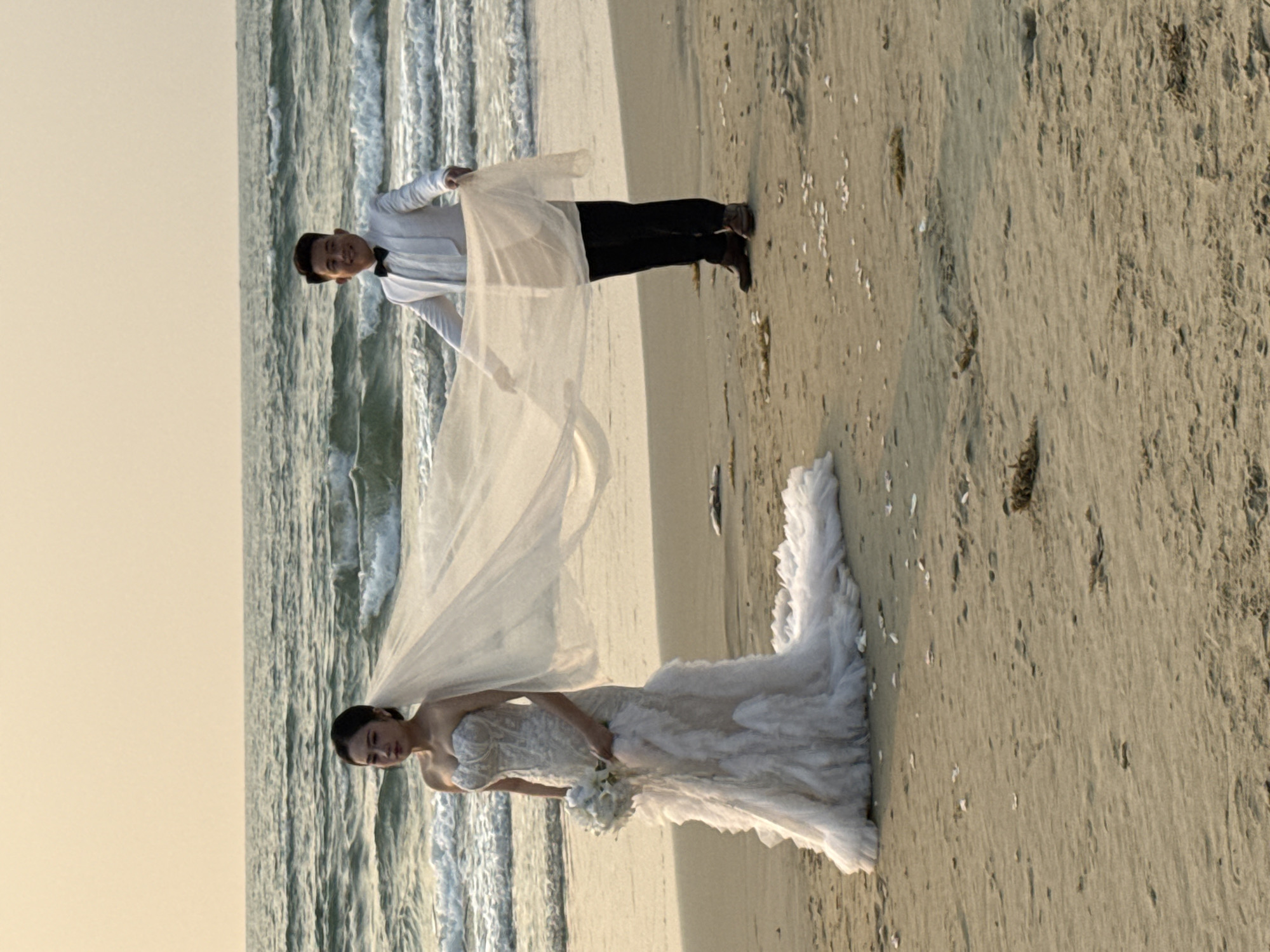 A wedding photo shows a groom holding the bride's veil on a beach in Đà Nẵng, Vietnam. The bride is wearing a white dress, and the groom is in a white shirt and black pants. The ocean is in the background.