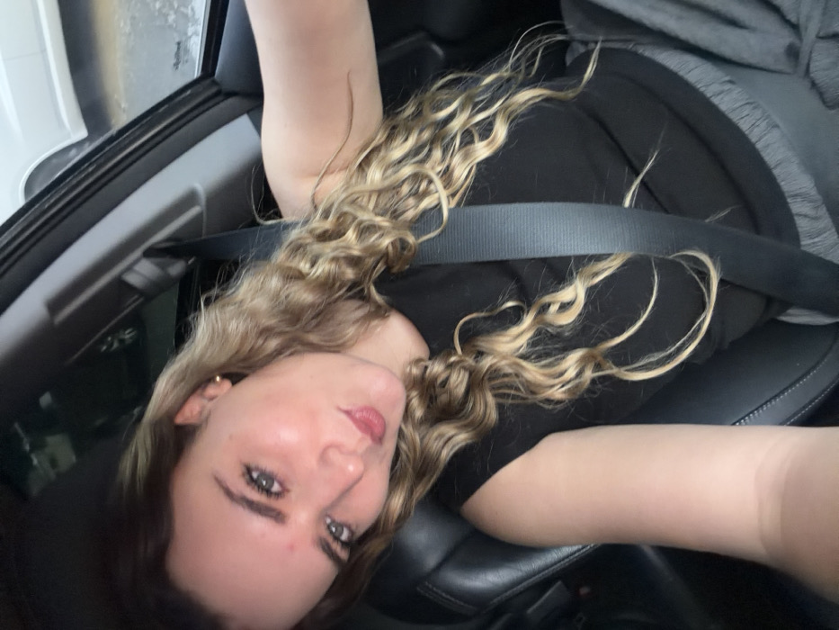 A single female individual is depicted in a selfie captured from within a vehicle. She has light-colored, wavy to curly hair and fair skin, with a small, spherical earring visible on her left earlobe. She is wearing a black sleeveless top. A dark-colored seatbelt diagonally crosses her torso, indicating she is secured in the passenger seat of the vehicle. Her head is tilted, and her facial expression includes slightly pursed lips while looking directly into the camera. The visible internal environment consists of dark upholstery, consistent with vehicle seating. Through the adjacent vehicle window, a partial view of a white vehicle and a wet, reflective ground surface is discernible. The precise location within the city of Puebla, Mexico, cannot be visually determined from the provided image data.