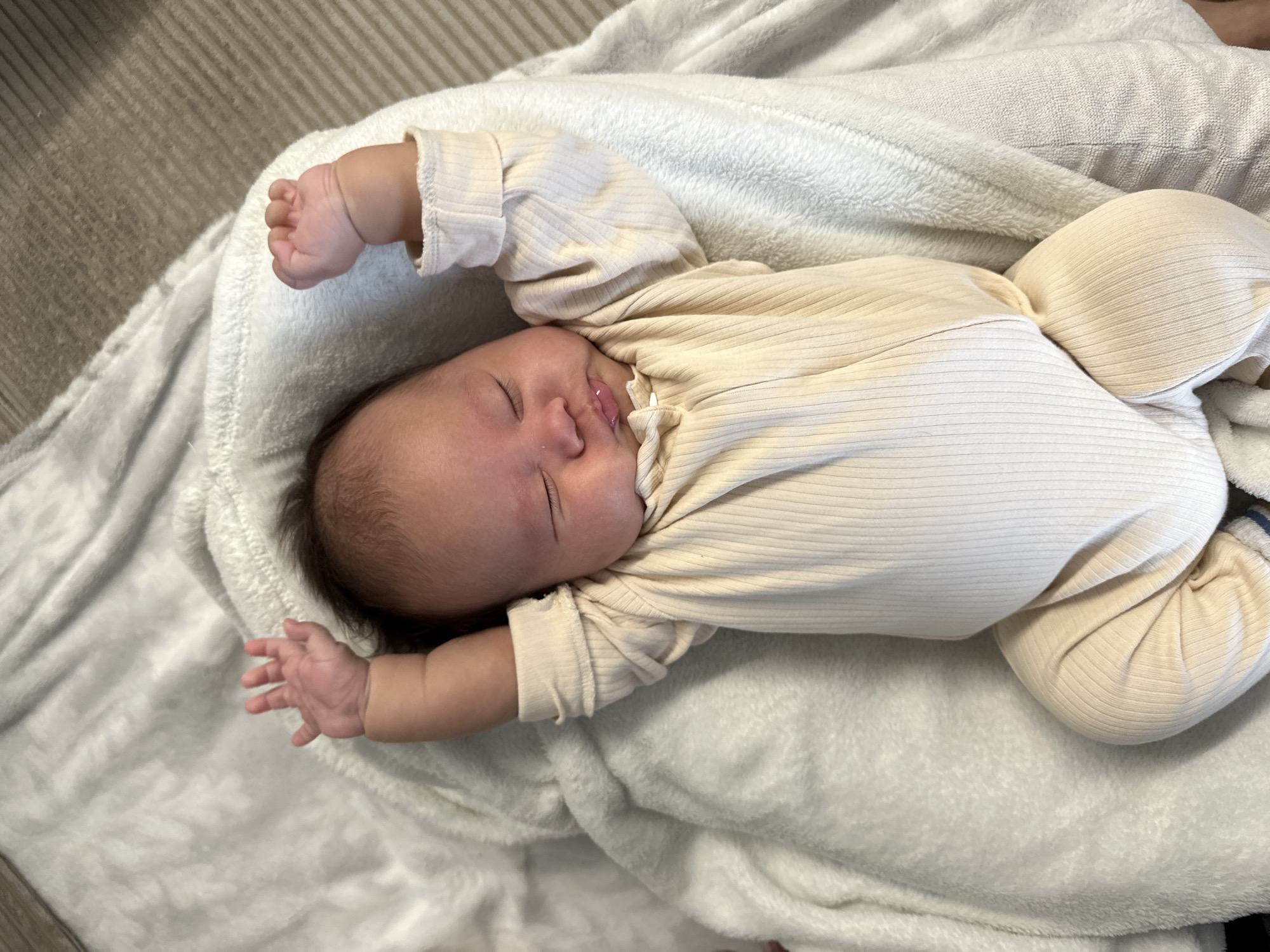 An infant is captured in a high-angle shot, sleeping peacefully on a soft, light-colored blanket. The baby, with dark hair, lies on its back, positioned slightly diagonally in the frame. Its eyes are closed, and its mouth is slightly open, indicating deep slumber. Both of the baby's arms are outstretched above its head, with relaxed, tiny hands visible.
The baby is dressed in a light cream or beige long-sleeved outfit, which features a distinct ribbed texture. Below the waist, the baby's bent legs are visible, also covered by the same ribbed material. The blanket directly beneath the baby is plush and appears to be white or a very light grey. In the background, to the left of the baby, a different surface with faint diagonal patterns is visible, suggesting an indoor setting like a floor or furniture. The lighting is soft and even, contributing to the tranquil atmosphere of the scene. No text is visible.
