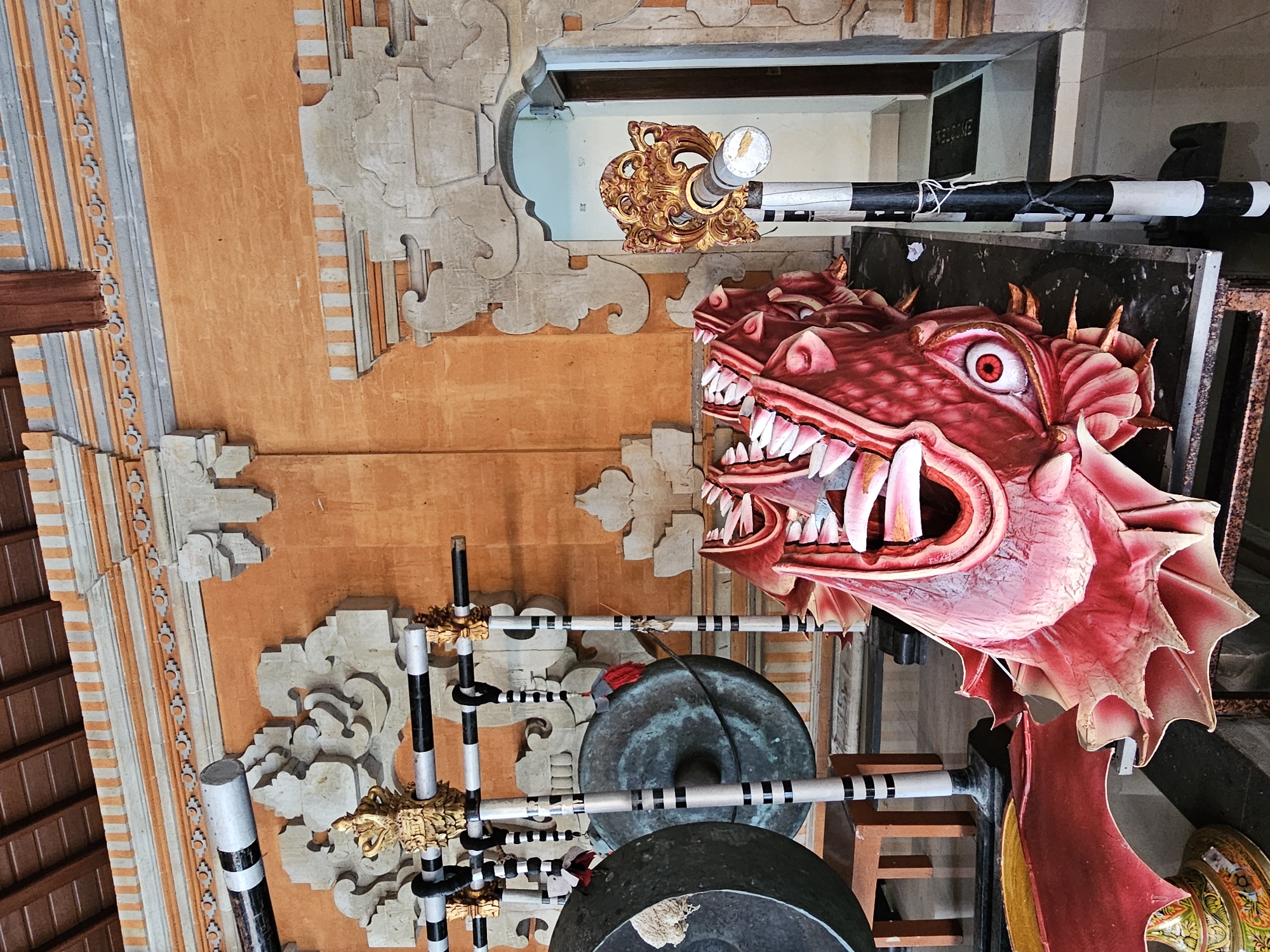 The image depicts the interior of a building in Denpasar, Indonesia, likely a temple or cultural center. In the foreground, a large, brightly colored dragon effigy, predominantly red and pink with a gaping mouth and prominent eyes, rests on a dark surface. To the left, two large gongs are suspended from a metal rack adorned with black and white striped poles and golden decorative elements. The walls of the building are painted a vibrant orange, with intricate stonework and decorative trim. In the background, an arched doorway leads to another room, with a welcome mat visible on the floor. The ceiling is constructed of dark wooden beams. The overall scene suggests a cultural performance or ceremonial setting, possibly related to Balinese traditions.
