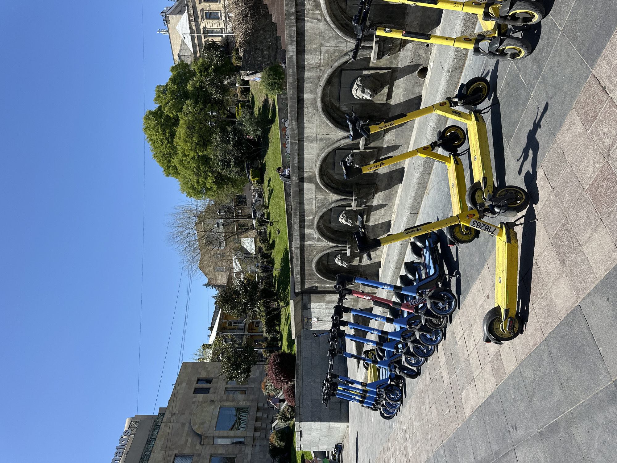 The image shows a row of electric scooters lined up on a paved sidewalk. The scooters are of two distinct brands: Yandex Go (yellow) and JET (blue with red accents). The background includes a concrete wall with circular, lion-headed fountains and a green, grassy area with trees. The location appears to be in Tbilisi, Georgia, based on the presence of Yandex Go scooters which are common in the city. A building is on the left side, and the sky is clear and blue. There are no visible people interacting with the scooters, however, there is at least one person sitting on the grass in the background.