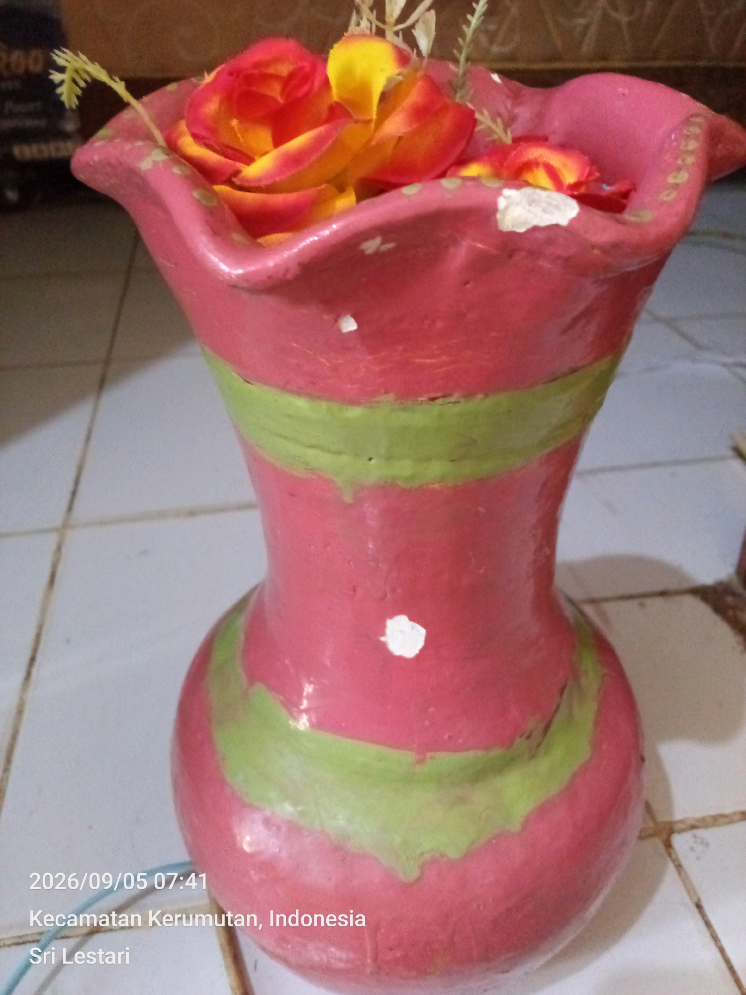 A close-up shot shows a pink vase with green bands, containing artificial red and yellow roses. The vase is chipped in a few places and has some white paint splatters. A small green plant stem extends from the top of the vase. The vase sits on a tiled floor. In the bottom left corner of the image, a timestamp "2026/09/05 07:41" and location information "Kecamatan Kerumutan, Indonesia Sri Lestari" are visible. The background is blurred, showing a wall with some objects.