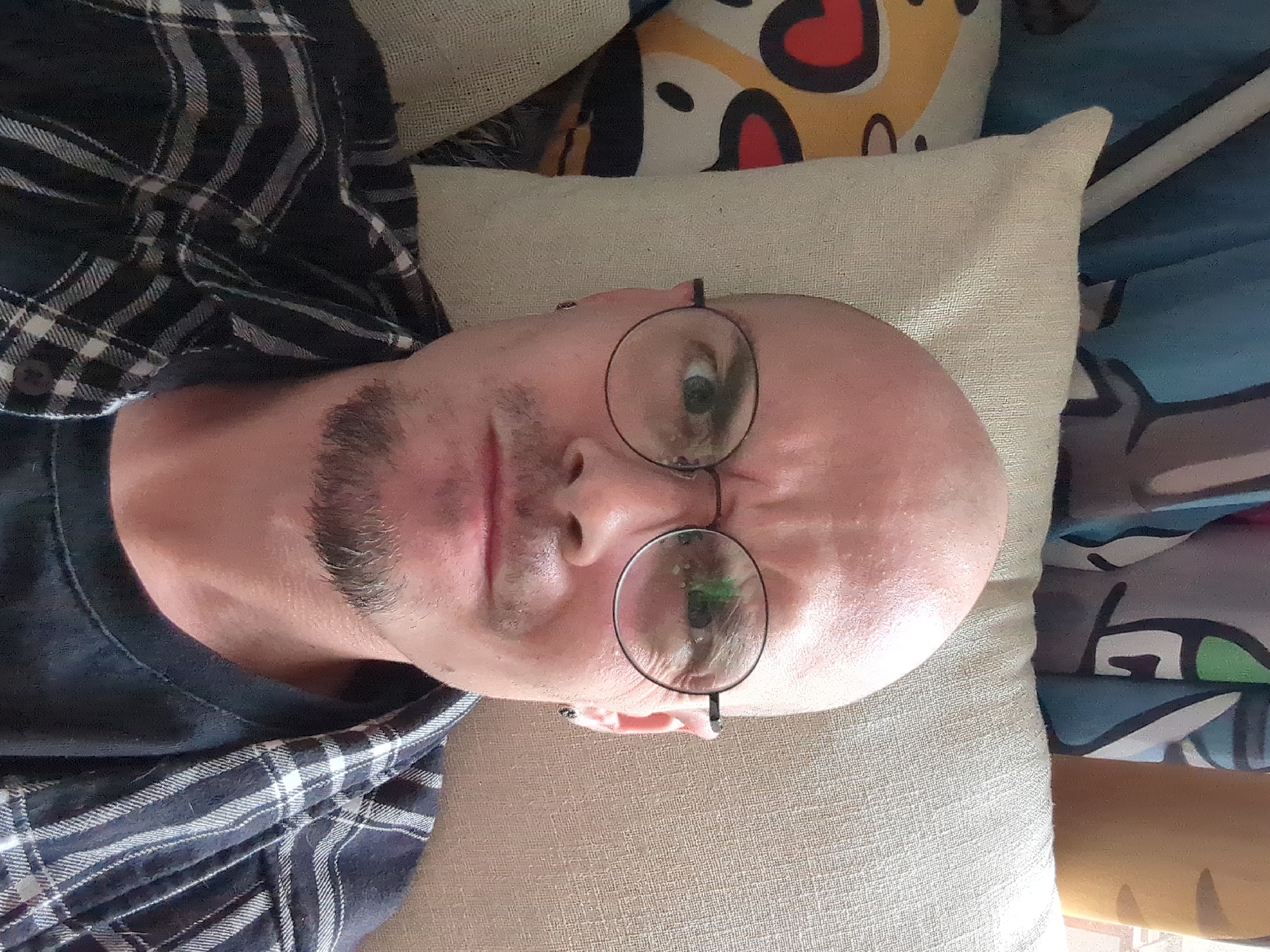 A close-up, rotated selfie depicts a bald Caucasian male wearing round-rimmed glasses and a dark blue and white plaid shirt. He has a goatee. He is lying down on a textured, beige pillow. Behind the pillow are colorful cushions, including one with a heart design and another with blue and green patterns. The location, Shunde District, China, cannot be verified based on the visual information alone.