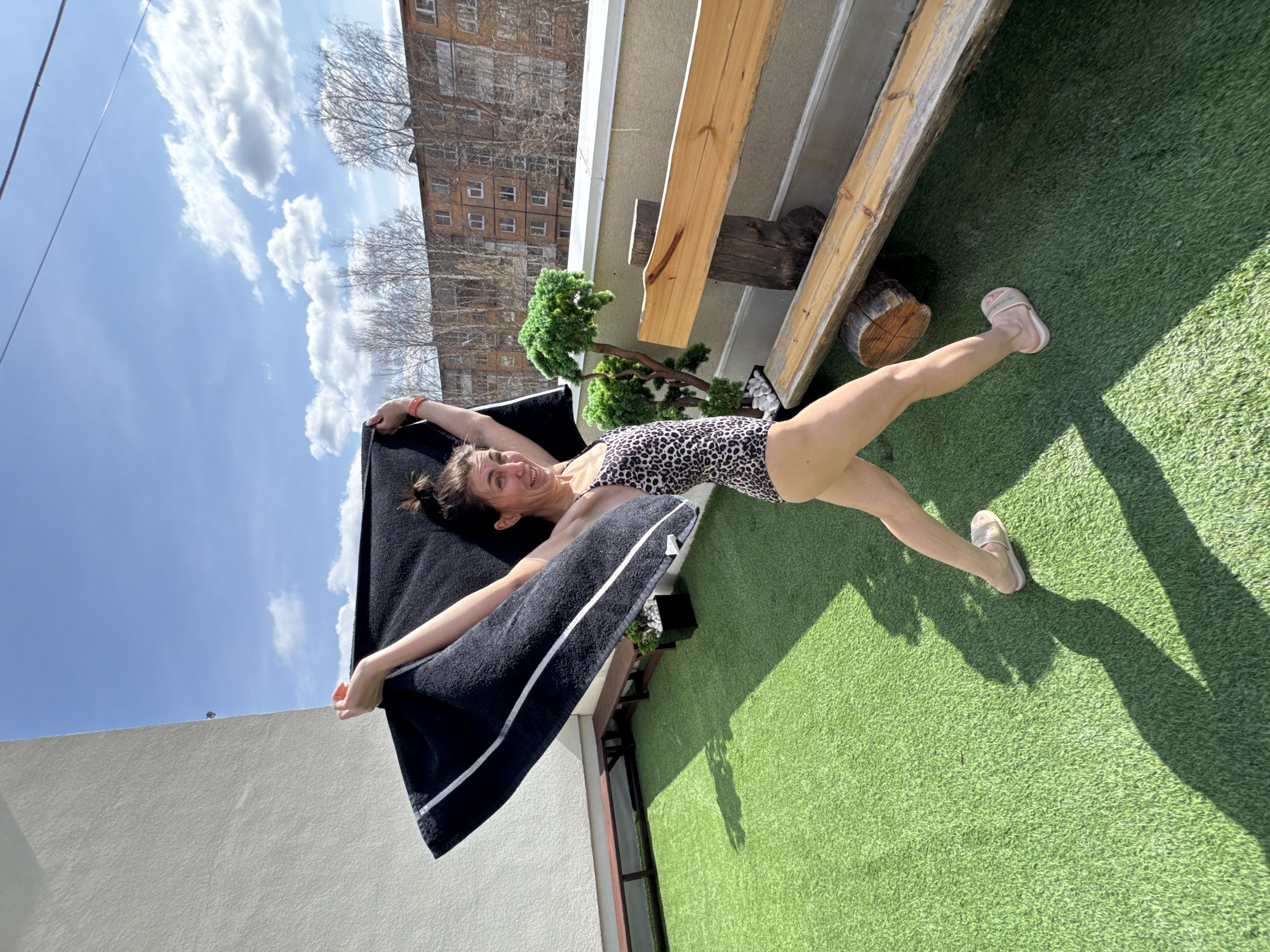 A woman in a leopard print swimsuit and pink sandals is lying on her back on a patch of artificial green turf on a balcony in Votkinsk, Russia. She is holding a black towel with a white stripe and looking at the camera with a smile. The balcony is furnished with a bench made of wooden planks and a small artificial tree. In the background, there is an apartment building, blue sky and scattered clouds. The weather appears sunny.