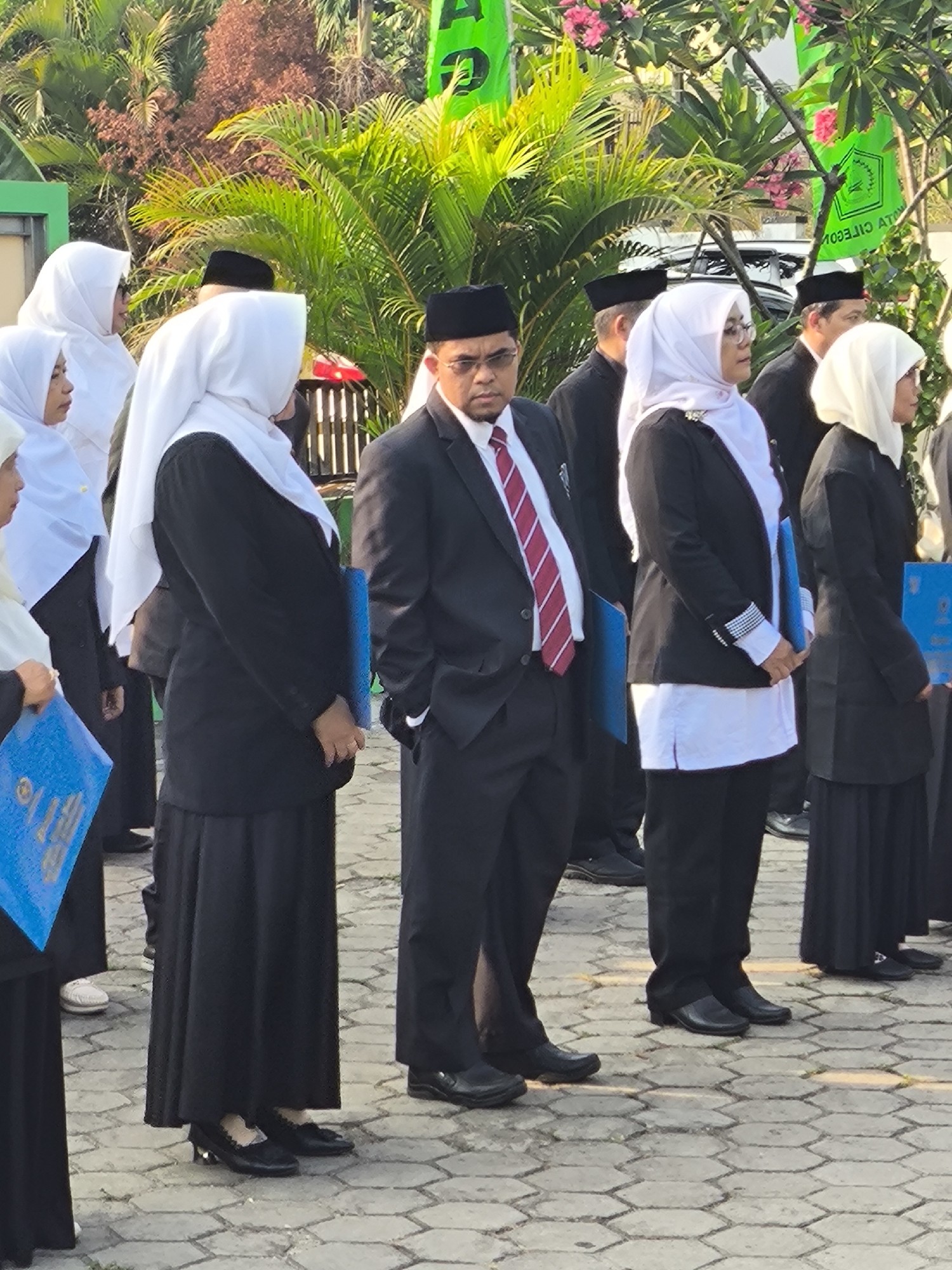 The image shows a group of people, mostly women wearing headscarves and black attire, standing outdoors on a paved walkway. Some are holding blue folders. In the background, there are lush green palm trees and pink flowers. Several green banners with text are visible. A man in a black suit and tie, also wearing a black cap, stands in the foreground, facing right. The overall scene suggests an outdoor event or ceremony. The presence of the banners with Indonesian text strongly suggests the location is in Indonesia. Considering the visual elements, particularly the foliage and architectural hints in the distance, it's plausible this scene is within a public space or institution in Cilegon, Indonesia.