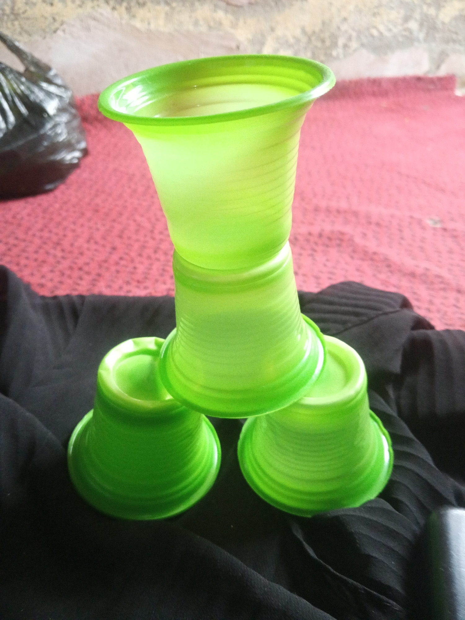 Four bright green plastic cups are stacked in a pyramid formation on a black cloth. Three cups form the base, with their openings facing upwards, and a fourth cup is placed on top, inverted. The cups have a ribbed texture. In the background, a red patterned cloth is visible, and further back, a crumpled black plastic bag. The scene appears to be outdoors, with a rough, textured wall or ground in the far background. The lighting suggests it is daytime. The specific location in Jalingo, Nigeria, is not discernible from the image alone.