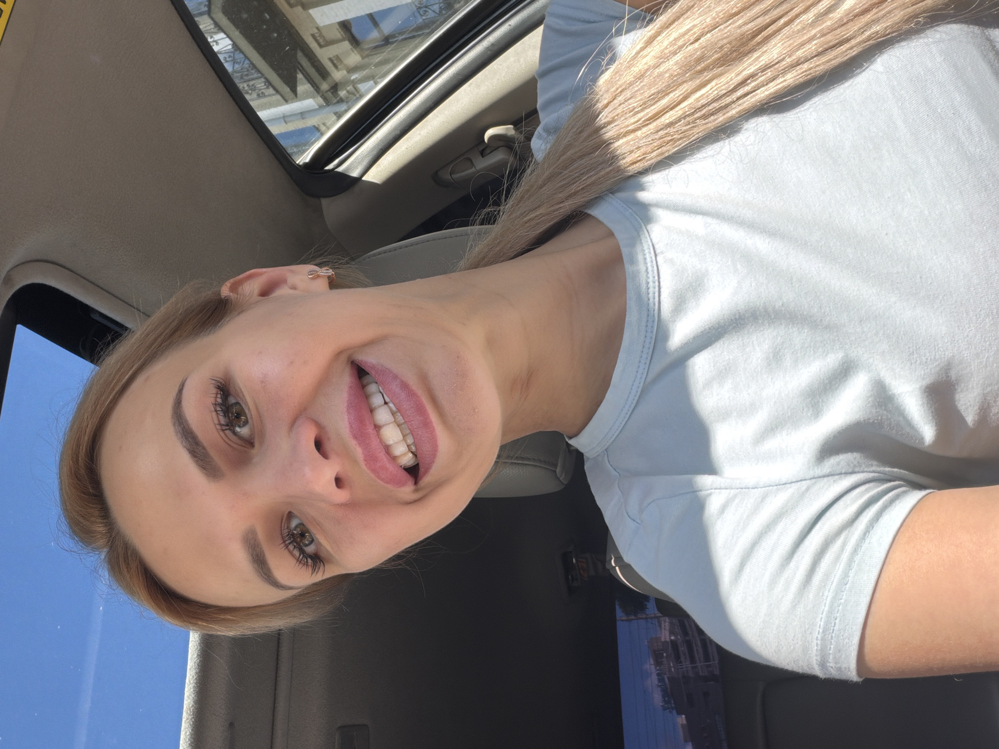 In a selfie taken from inside a car, a young woman with blonde hair pulled back in a ponytail smiles with her mouth open, revealing her teeth. She is wearing a light blue t-shirt and a small, rose gold hoop earring in her left ear. The car's sunroof is open, allowing a view of the clear blue sky. The background shows the interior of the car, including a grey leather seat and the passenger side window, through which a building with balconies and decorative railings is visible. The scene appears to be set on a sunny day in the city of Kyiv, Ukraine.