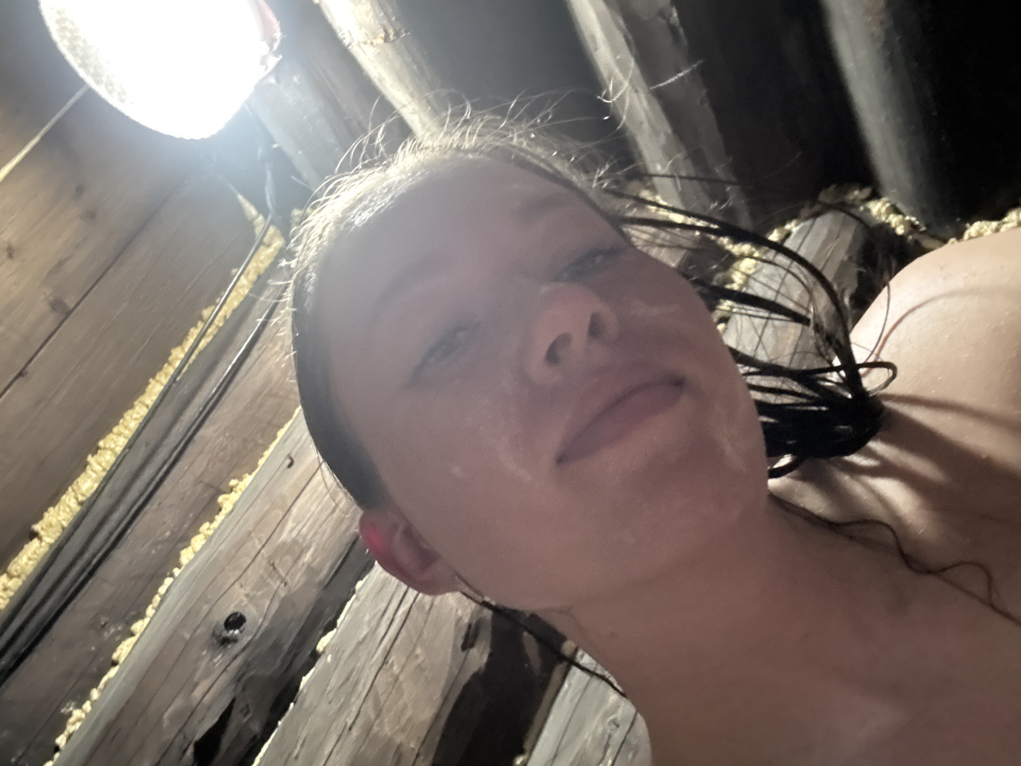 A close-up, low-angle photograph shows a person with light skin and wet hair, possibly female, under a wooden structure. The structure appears to be an attic or similar confined space, with exposed wooden beams and some sort of insulation or sealant between the beams. A light source illuminates the upper left corner of the image. The location is Ферма, Russia.