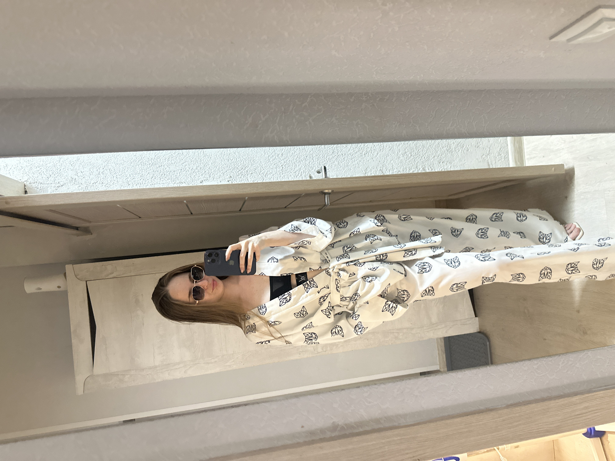 A young woman with long, light brown hair and sunglasses stands in a room, taking a mirror selfie. She is wearing a white satin robe with a black cat print and a black bra. The robe is tied at the waist with a matching belt. She is holding a dark-colored smartphone in her right hand. The room has a light-colored door on the right and a tall, light-colored wardrobe on the left. The floor is light wood. The lighting appears to be natural, coming from an unseen source.