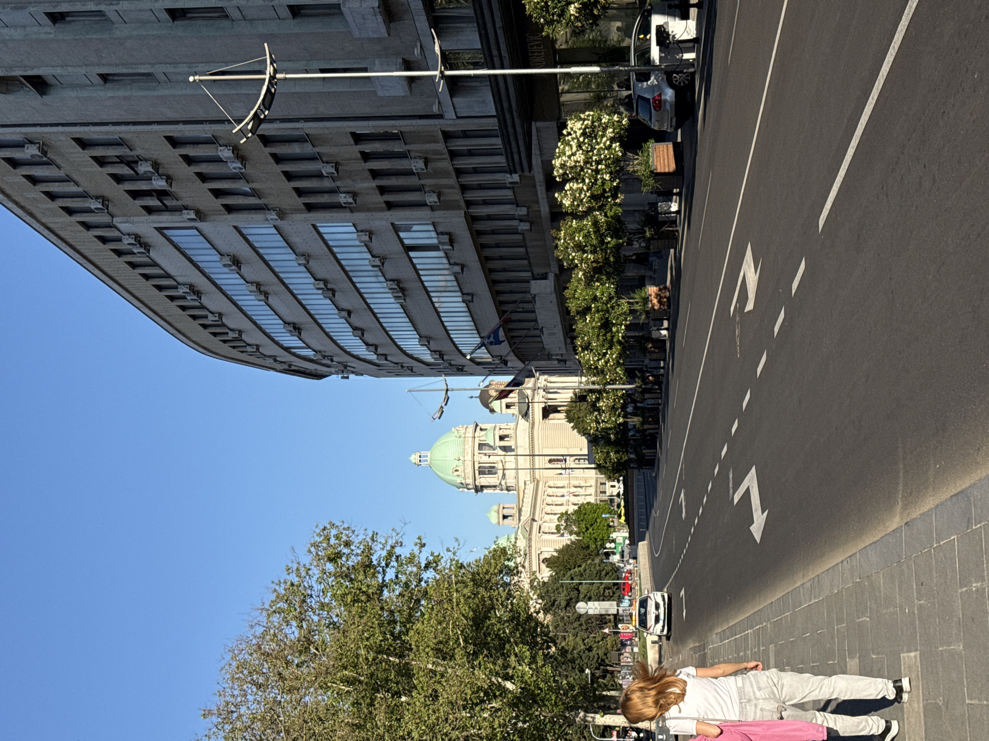 The image shows a street scene in Belgrade, Serbia, featuring the National Assembly Building with its distinctive green dome. A modern, curved building stands on the left side of the road. A person is lying on the sidewalk in the foreground, partly obscured by trees. A road with painted direction arrows runs between the sidewalk and the buildings. Cars are visible on the road. The sky is blue and clear.