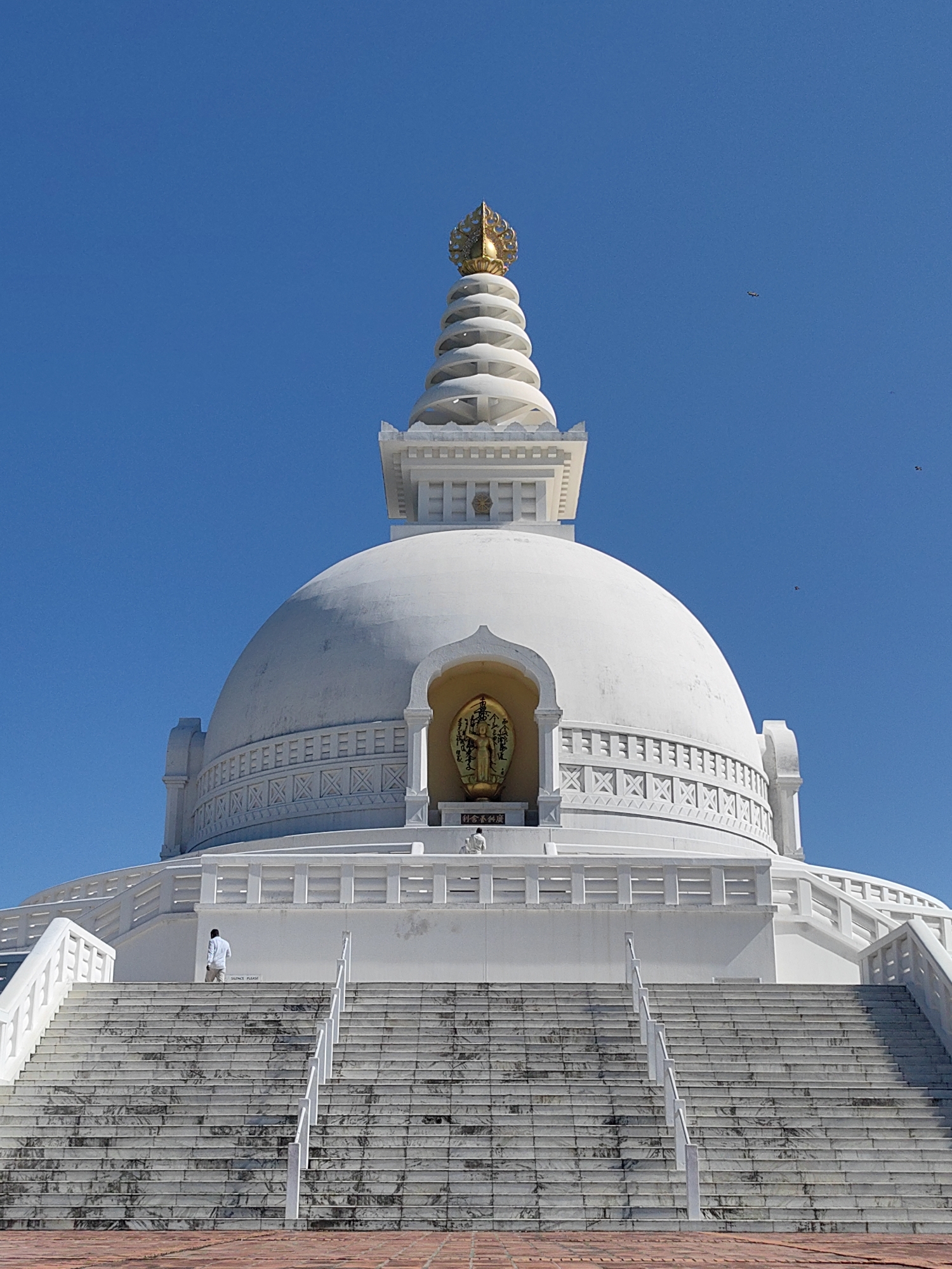 A bright, low-angle, full-frame shot captures the Peace Pagoda in Lumbini, Nepal, against a clear, bright blue sky. The white stupa, topped with a golden spire, dominates the frame, with a large white dome forming its base. A gold statue of Buddha is visible in an alcove on the dome. Wide, white marble stairs lead up to the stupa, with a few individuals ascending. A man in a white shirt and tan pants is halfway up the stairs, walking towards the stupa. Another person, dressed in white, is seated near the top of the stairs. Small birds fly in the distance. The foreground consists of red brick paving.