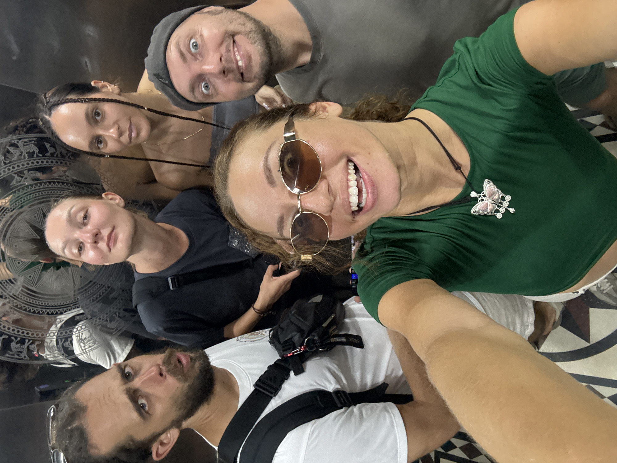 This is an upward-angled group selfie featuring five adults in a highly reflective indoor space, possibly an elevator or mirrored room.
In the foreground, a woman with curly brown hair, wearing round sunglasses and a deep green top, smiles widely, revealing her teeth. A prominent silver and pink butterfly necklace with clear stone accents adorns her chest. Her outstretched arm suggests she is holding the camera. Directly above her, a man with a short beard and a grey beanie smiles towards the lens. To the mid-left, a woman with braided dark hair looks at the camera with a calm expression.
In the bottom left of the frame, a bearded man wearing a white t-shirt and a black harness or backpack with an "adidas" logo visible on one strap looks upward with a slightly surprised or intense expression. Another woman, wearing a black t-shirt, is partially visible in the mid-left background, also looking towards the camera.
The reflective surfaces behind the subjects display distorted reflections of the individuals and a distinct circular pattern on what appears to be a floor or decorative panel. This ornate design features intricate geometric shapes and stylized animal figures, characteristic of traditional Southeast Asian art. The overall atmosphere is casual and cheerful, with most subjects smiling. The indoor lighting does not provide specific cues about the time of day or weather. The location context of Hà Giang 1 Ward, Vietnam, aligns with the potential cultural motifs seen in the background pattern.