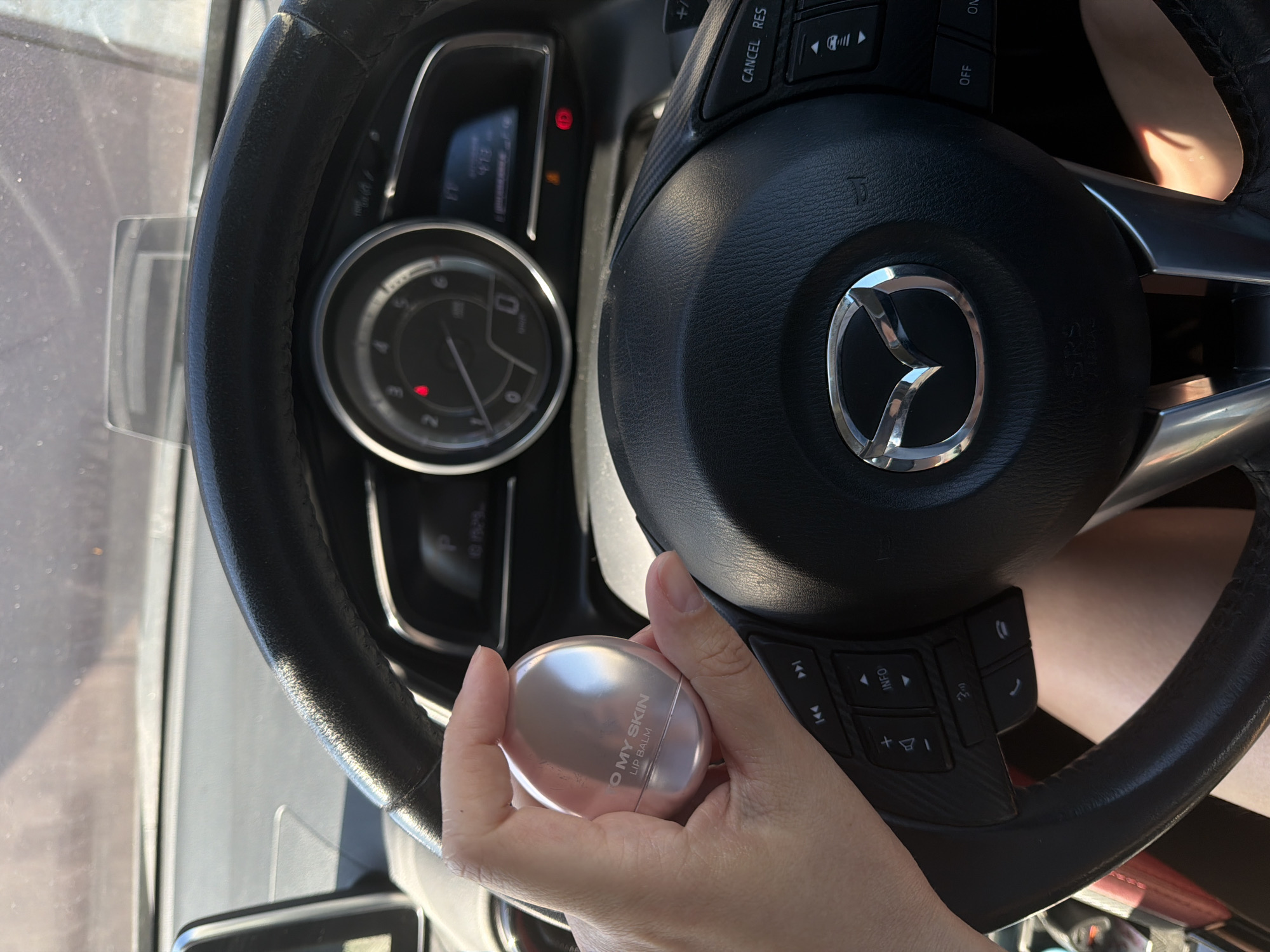 An overhead, close-up shot shows a person's hand holding a small, round, silver-colored lip balm container in a car. The lip balm has "TO MY SKIN LIP BALM" printed on it. The person's hand is positioned near the steering wheel of the car. The steering wheel has a Mazda logo in the center and various buttons on the right side, including "CANCEL RES". The dashboard of the car is visible in the background, with a speedometer and digital display. The lighting suggests it is daytime and the car is parked or stopped, as indicated by the sunlight reflecting off surfaces. The image does not provide any information about the location being in the city of Chaikovsky, Russia.