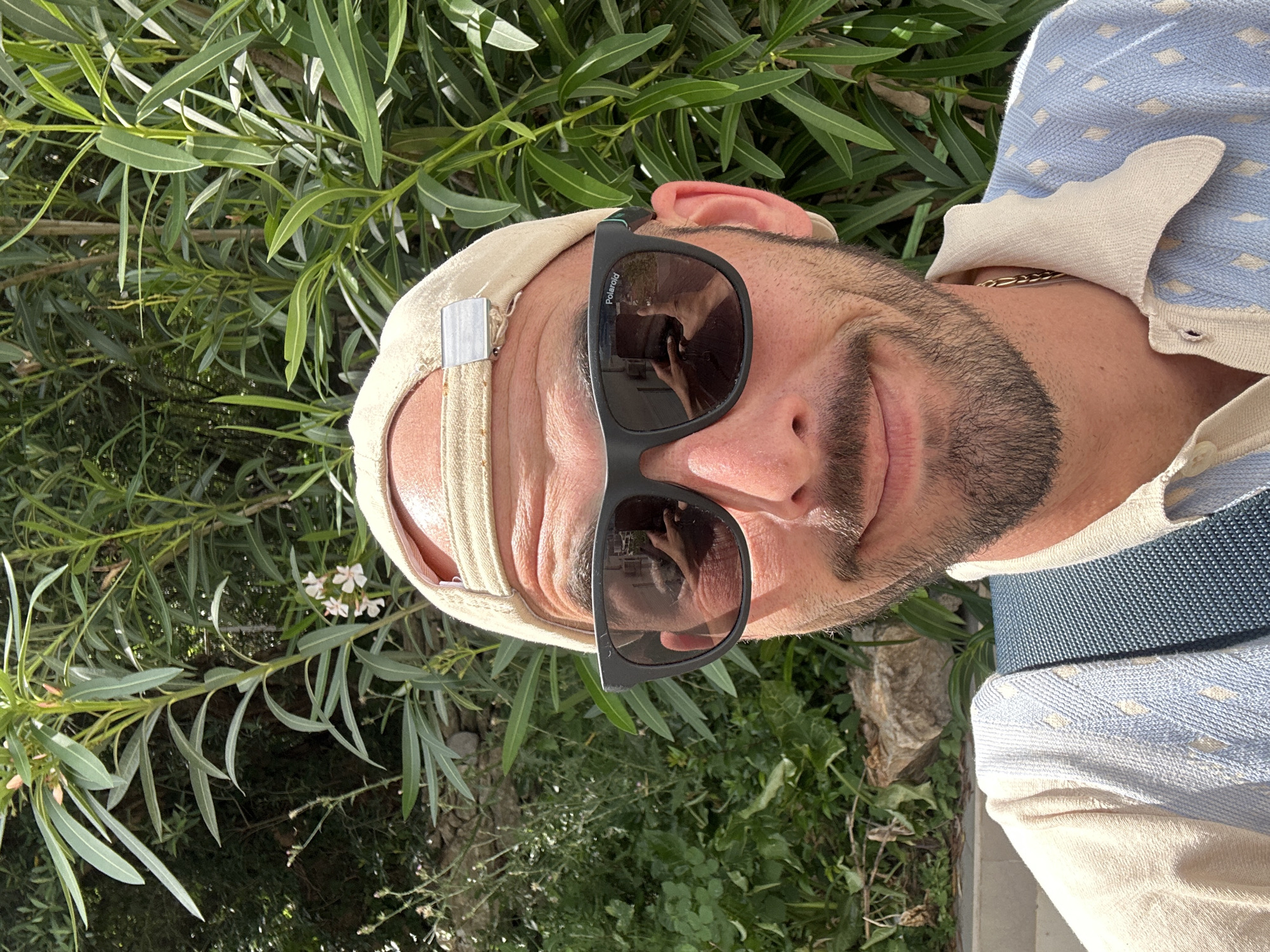 A man is centered in the image, with greenery surrounding him. He wears a beige baseball cap backwards, black Polaroid sunglasses, and a light blue and beige button-down shirt with a gold chain visible at the neck. The man is looking at the camera with a slight smile. The foliage consists of green leaves. The location is likely Budva, Montenegro, based on the light conditions and vegetation, but no landmarks are visible to confirm this.