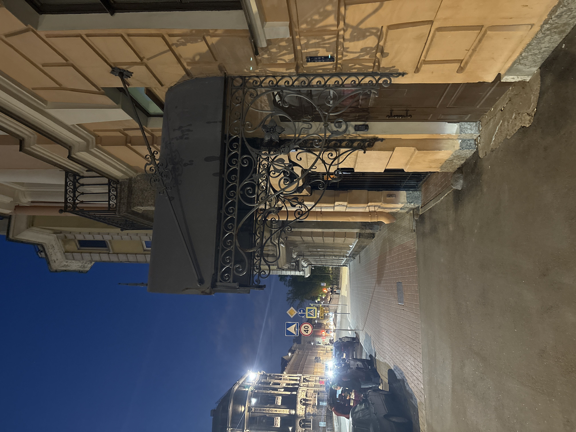 The image depicts a street scene in Saint Petersburg, Russia, at dusk or night. The foreground features a building with ornate wrought-iron details above a doorway, situated along a brick-paved street. The street extends into the distance, illuminated by streetlights. Various traffic signs are visible, including a speed limit sign with the number "40." Vehicles, primarily scooters, are parked along the side of the street. The sky is a gradient of blue, indicating the transition from day to night. The buildings along the street exhibit architectural details typical of Saint Petersburg, with multiple stories and decorative facades. There are people, but they are hard to identify due to distance and darkness.