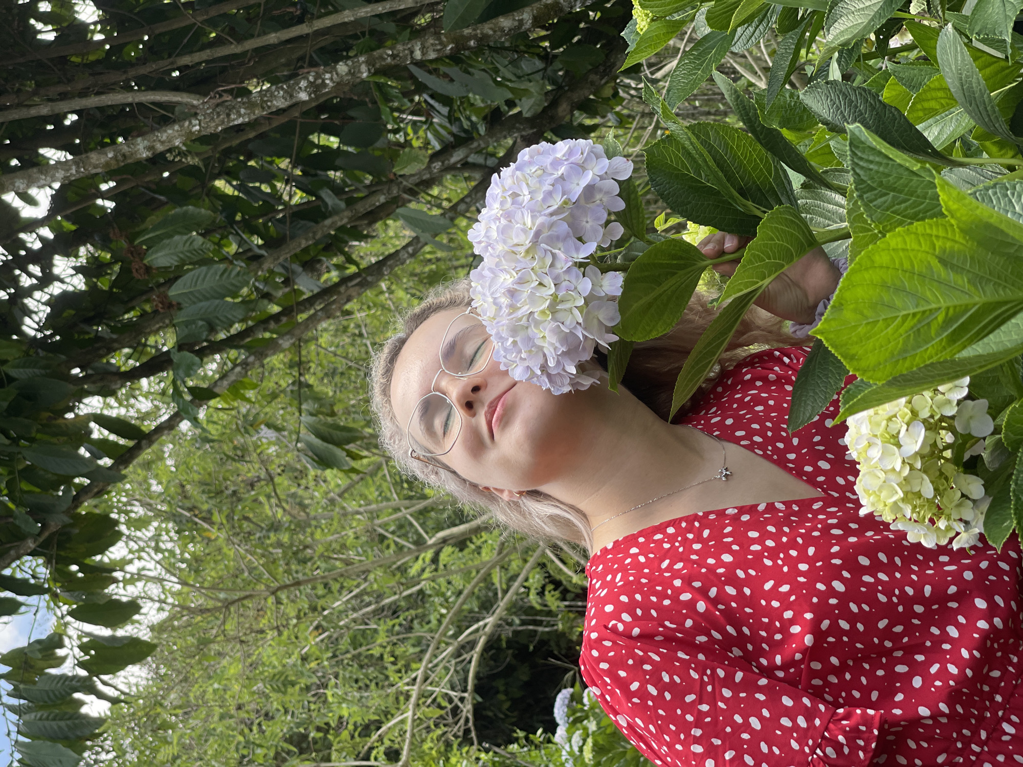 A young woman stands in an outdoor setting surrounded by verdant foliage and flowers, possibly in Da Lat, Vietnam. She wears glasses and a red dress with white polka dots, with a silver necklace visible at her throat. A large, light purple hydrangea bloom is positioned near her face, obscuring one eye. Her eyes are closed, and her head is tilted slightly upwards. Behind her and around her are various plants and trees, creating a lush background. The image is taken from a lower angle, looking up at the subject. In the periphery, additional hydrangea blooms can be seen.