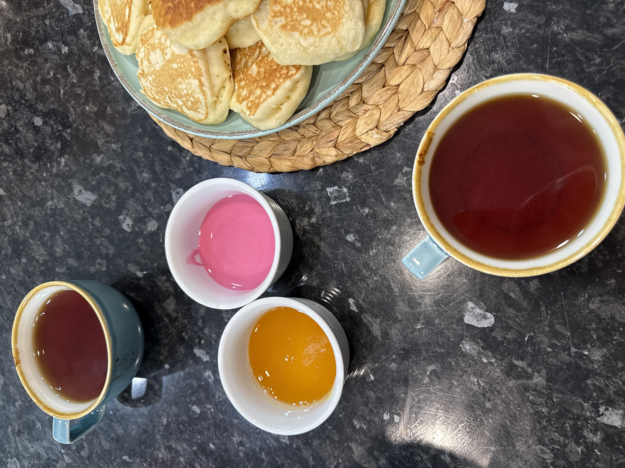 An overhead shot shows a dark, speckled countertop with two mugs of tea, two small white bowls, and a plate of pancakes. One mug, teal with a tan rim, is in the upper left corner. The second mug, white with a tan rim and a teal handle, is in the lower right corner, filled with dark amber liquid. To the left of the second mug are two small white bowls: one containing a bright orange liquid, presumably honey or jam, and the other containing a bright pink liquid, possibly a jam or a topping. To the right of the bowls is a light blue plate filled with golden-brown pancakes, piled on top of each other and resting on a round, woven placemat. The scene is well-lit, with reflections visible on the countertop. There are no people or animals present.