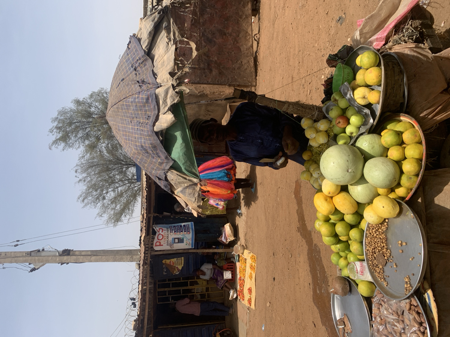 This image captures a bustling open-air market scene in Rafin Par, Nigeria, under a bright, clear sky, suggesting midday or early afternoon due to the strong sunlight and shadows.
In the right foreground, a fruit vendor stands behind a colorful stall. The vendor, a man dressed in a dark blue tunic, is partially visible beneath a makeshift awning made of patterned fabric and a green tarp. The stall is laden with a vibrant assortment of fresh produce displayed in metal trays and woven baskets on the dusty ground. Prominently featured are several pale green watermelons, surrounded by piles of yellow fruits (possibly mangoes or papayas), smaller green fruits (limes or unripe citrus), and numerous small, bright yellow-orange citrus fruits. One tray also holds a collection of small, light brown nuts or legumes, while another contains what appears to be dried fish and a coconut.
Behind the fruit stall, several simple market structures or shops with open fronts line what appears to be a dirt path. One of these structures features a blue and white sign with the text "POS AVAILABLE HERE," indicating the availability of point-of-sale services. Other individuals are sparsely visible in the background near these structures. On the left side of the frame, a tall, concrete utility pole rises against the clear sky, with the sparse foliage of a tree visible further in the distance. The ground throughout the market area is unpaved and dusty.
