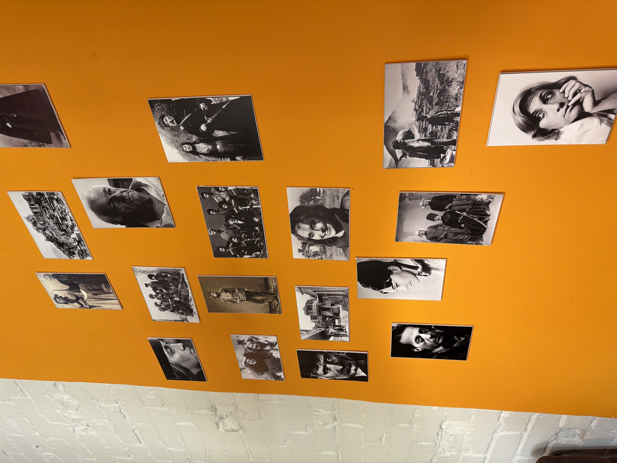 The image shows a brightly lit orange wall adorned with approximately 18 black and white photographs of varying sizes. The photos include portraits of people, groups of individuals in historical attire, landscapes, and urban scenes. The photographs are arranged in a somewhat haphazard manner. Below the orange wall, there is a section of wall painted with white bricks. The location appears to be indoors. There is no clear evidence in the image to confirm it is located in Moscow, Russia.