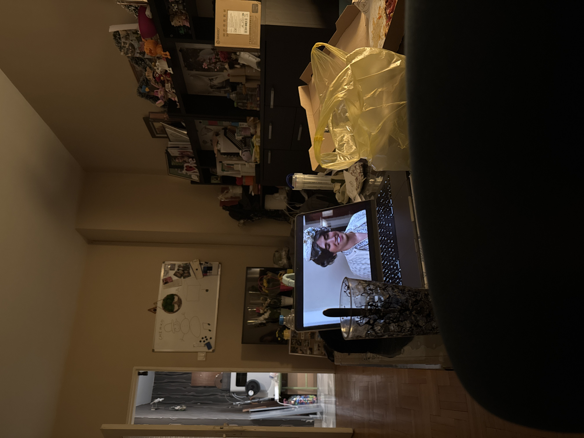 The image shows a cluttered interior scene. In the foreground, a laptop displays a woman with dark hair and a white hat, possibly on a video call. A patterned glass with a spoon is visible near the laptop. The background includes a bookcase filled with various items, boxes, and a yellow plastic bag. A whiteboard with drawings and notes is mounted on the wall. Through an open doorway, a glimpse of another room with a dark wavy design on the floor is visible. The location is likely Sofia, Bulgaria, as the prompt requests, but this is not verifiable from the image itself. The scene suggests a personal living space.