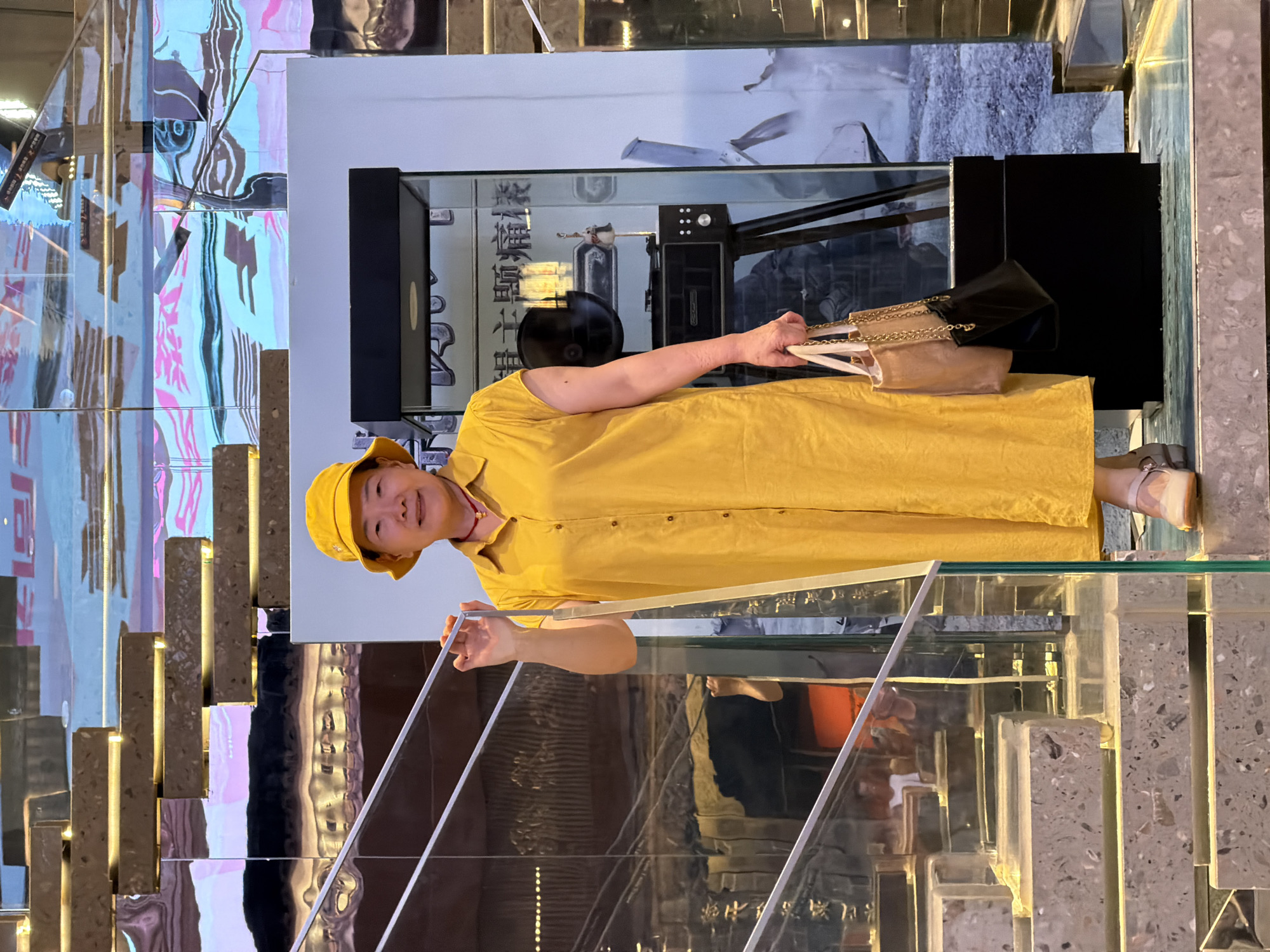 A woman stands on a glass staircase in Yuzhong District, China, holding two handbags. She is wearing a yellow hat and a yellow dress. The staircase is made of glass and concrete. In the background, there is a display case with various objects. The overall environment appears to be an indoor public space with reflective surfaces and artistic displays.