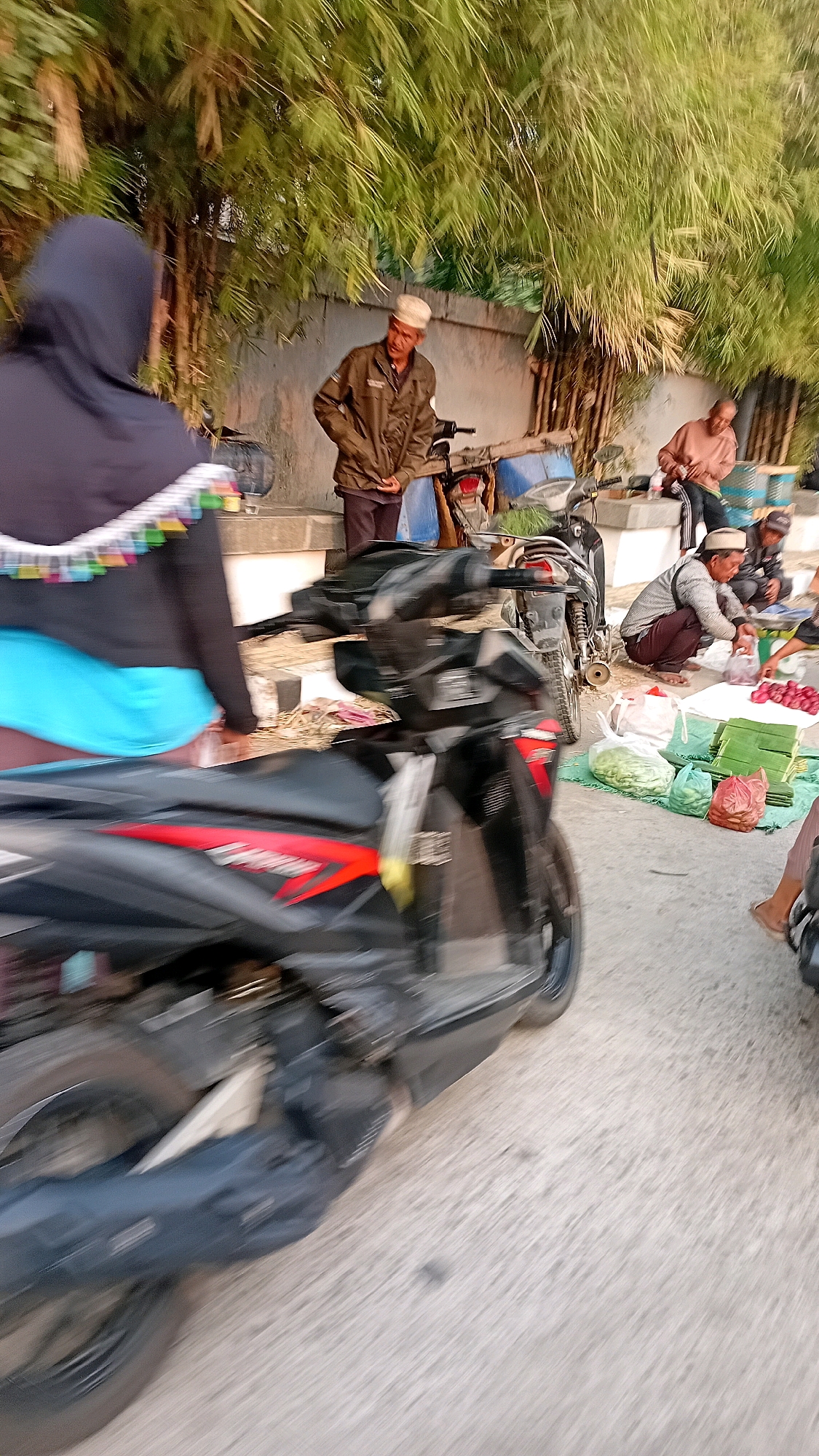 This is a motion-blurred, eye-level, medium shot of a street market scene, likely in Jambi, Indonesia, during daylight hours. In the foreground, a black and red motorcycle is in motion, with its rear wheel and body significantly blurred. To the left, a person wearing a dark hijab and a blue garment is partially visible, facing away from the camera.
In the background, several individuals are engaged in activities related to market sales. A man in a brown jacket and a white cap stands near a motorcycle. To his right, another man, also wearing a cap, is seated and appears to be interacting with produce laid out on green banana leaves and in plastic bags. These produce items include red onions and possibly other vegetables. Other individuals are further in the background, with one person in a light-colored shirt seated behind a stall. The scene is set against a backdrop of a textured wall and dense bamboo foliage. The ground surface appears to be paved.
