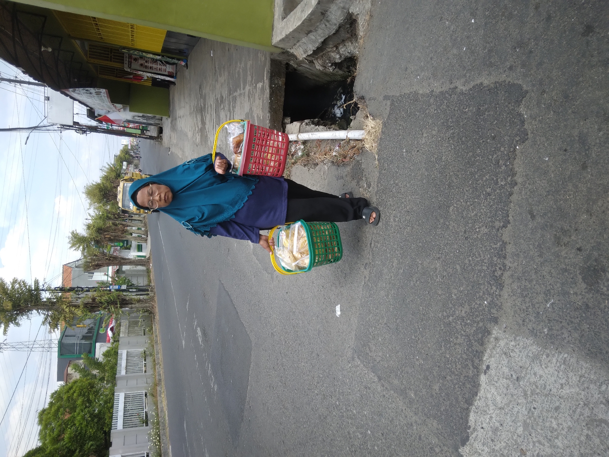 A woman wearing a teal hijab and glasses walks on a sidewalk beside a street in Purwokerto Wetan, Indonesia. She is carrying two baskets filled with food items, one green and one red. The street is paved and appears to be a commercial area with buildings and trees lining one side. The woman is walking away from the viewer, towards the right of the frame.