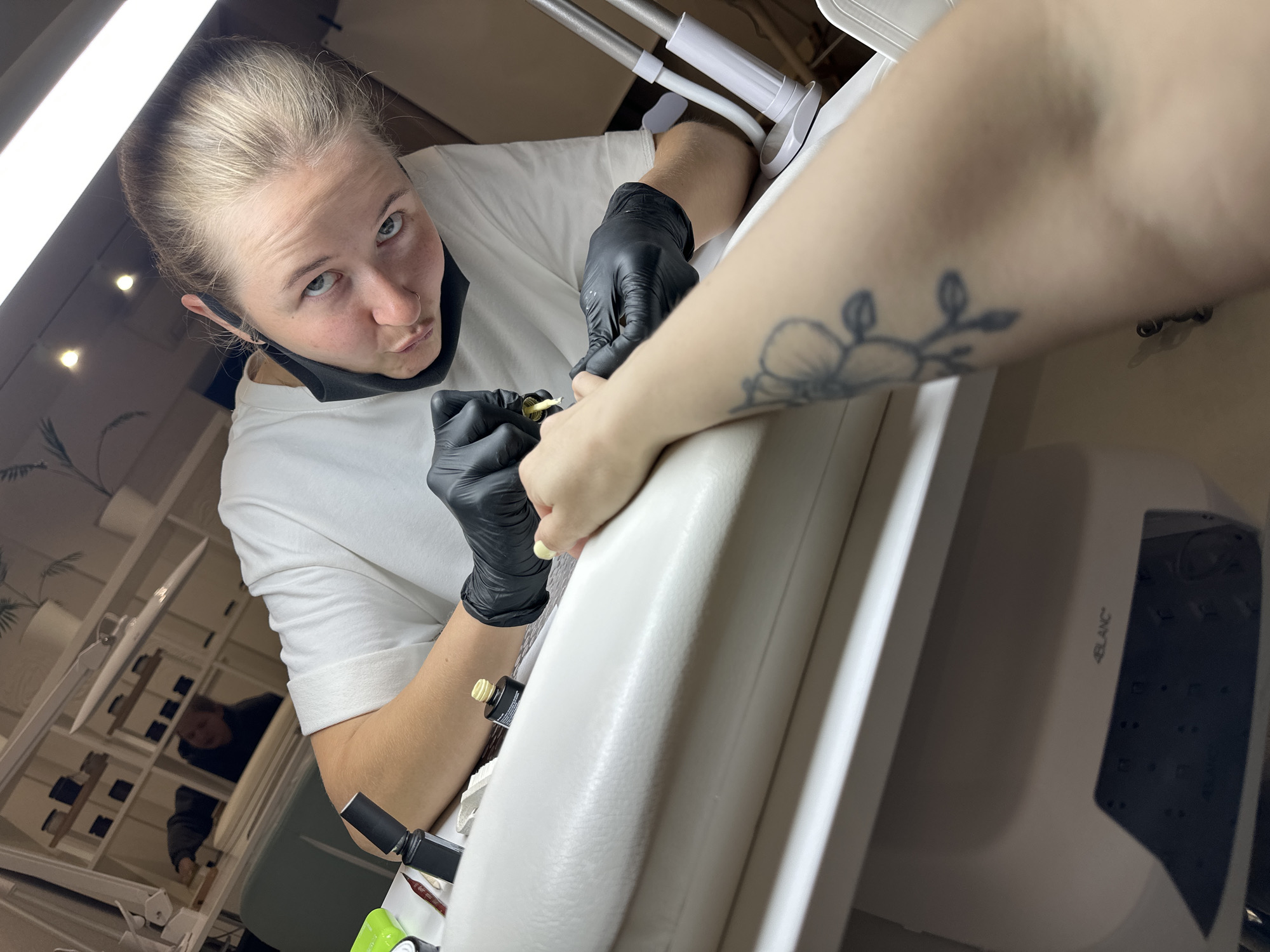 A close-up, first-person perspective photograph, taken in a nail salon in Perm, Russia, captures a female nail technician applying nail polish to a client's hand. The technician, with light-colored hair pulled back, blue eyes, a nose piercing, and a black face mask lowered to her chin, wears a white t-shirt and black disposable gloves. She is shown applying a light-colored polish to the client's fingernail using a small brush from a yellow-capped bottle, while looking directly at the camera with a slight pout.
Only the client's right arm and hand are visible in the foreground, resting on a white cushioned armrest. The client's inner forearm displays a faded floral tattoo, and the fingers show freshly applied light, creamy nail polish. The white workstation holds various nail products, including several black nail polish bottles, a green container, and small tools. An adjustable task lamp illuminates the client's hand, supplemented by a long, overhead LED light fixture.
The background, largely reflected in a glossy surface behind the technician, shows a blurred figure of another individual, possibly male and wearing dark clothing, seated at a desk. White shelving units with potted plants are also discernible. Under the main table, a piece of equipment marked "4BLANC" is partially visible. The overall environment appears clean, well-lit, and professionally maintained.