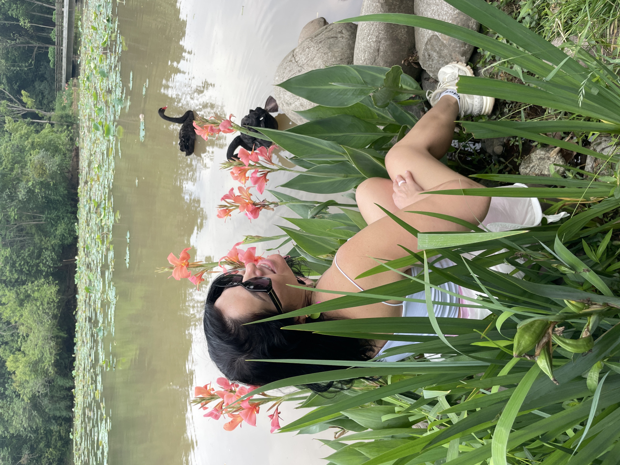 The image captures a woman lying amidst tall green foliage and vibrant orange flowers, with her head near the edge of a murky pond. She wears a striped top, white shorts, and white sneakers, and sports sunglasses. Two black swans are visible in the pond near her head, with numerous lily pads covering the water's surface further out. In the background, across the pond, a forested area and a bridge structure are seen. The location is Chenghua District, China.