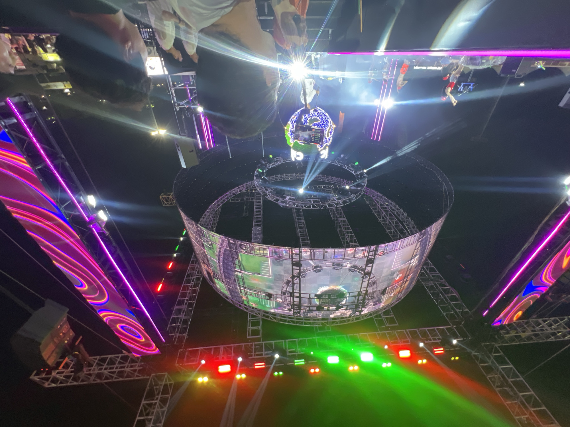 The image depicts an outdoor night concert at an unspecified location in Daegu, South Korea. A large spherical stage structure, adorned with lights and screens, dominates the center of the frame. This spherical stage is surrounded by complex metal scaffolding and further illuminated by colorful stage lights, including vibrant green, red, and purple beams. Several large, vertical LED screens displaying abstract, colorful patterns are positioned on either side of the main stage. On the main stage, performers are visible, some appearing to be dancing and interacting with the audience. A crowd of spectators is gathered in the foreground, facing the stage, with some individuals partially visible in silhouette. The overall atmosphere is energetic and festive, indicative of a large-scale entertainment event.