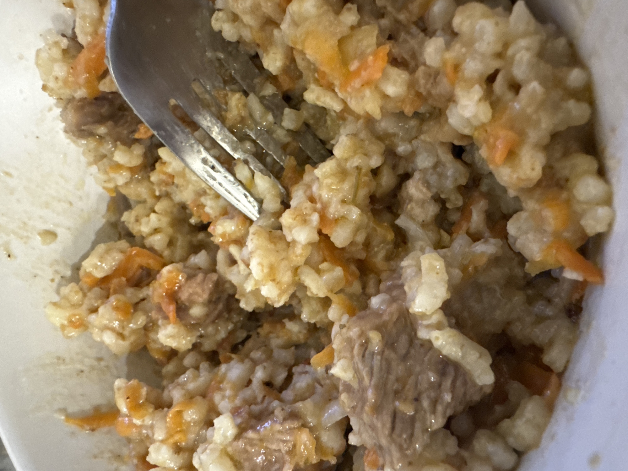 A close-up, overhead shot shows a bowl of pilaf. The dish contains rice, shredded carrots, and chunks of meat. A metal fork is visible, partially submerged in the pilaf, suggesting it is being eaten. The lighting is dim, and the bowl appears to be white porcelain. The setting is a domestic environment, likely a home kitchen. There is no visible connection to the city of Saratov, Russia.