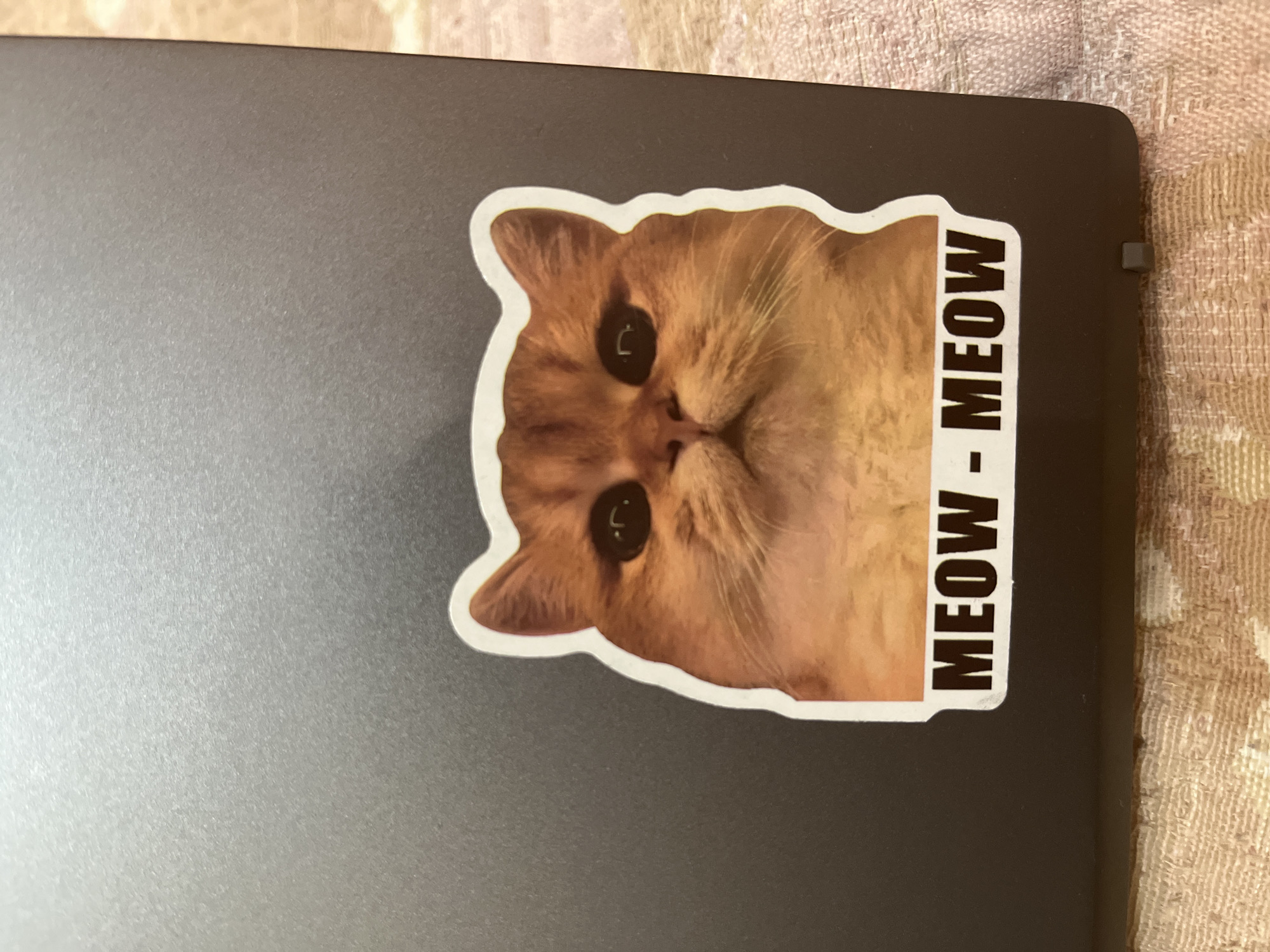 The image shows a close-up of a sticker of a ginger cat's face on a dark gray surface, likely a laptop. The cat has large, dark eyes and a slightly upturned mouth. Below the cat's face, the words "MEOW - MEOW" are printed in white capital letters on a black background. The surface the sticker is on appears to be textured and is resting on a light-colored, patterned fabric. There are no discernable landmarks or environmental cues that would indicate a specific location within Stara Zagora, Bulgaria.
