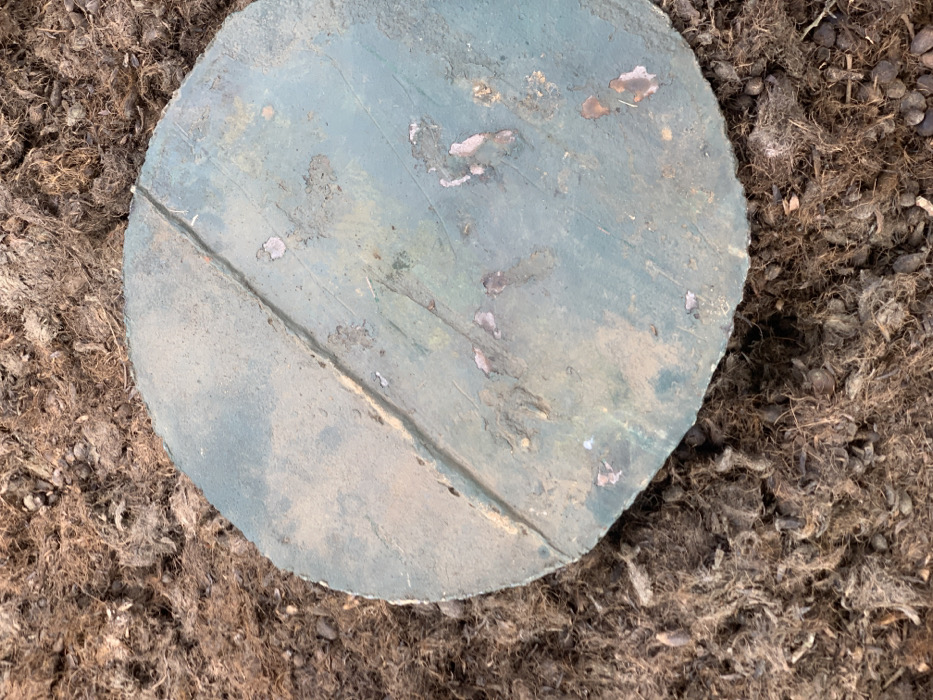 The image shows a circular, weathered, green concrete cover, partially resting on top of a bed of brown, fibrous, mulch. The cover has a large crack running through its center, with a few small, faded orange and pink patches scattered across its surface. There are no visible people, animals, or plants in the image. The location is Egbeda, Nigeria, based on coordinates (latitude: 7.119, longitude: 5.362).