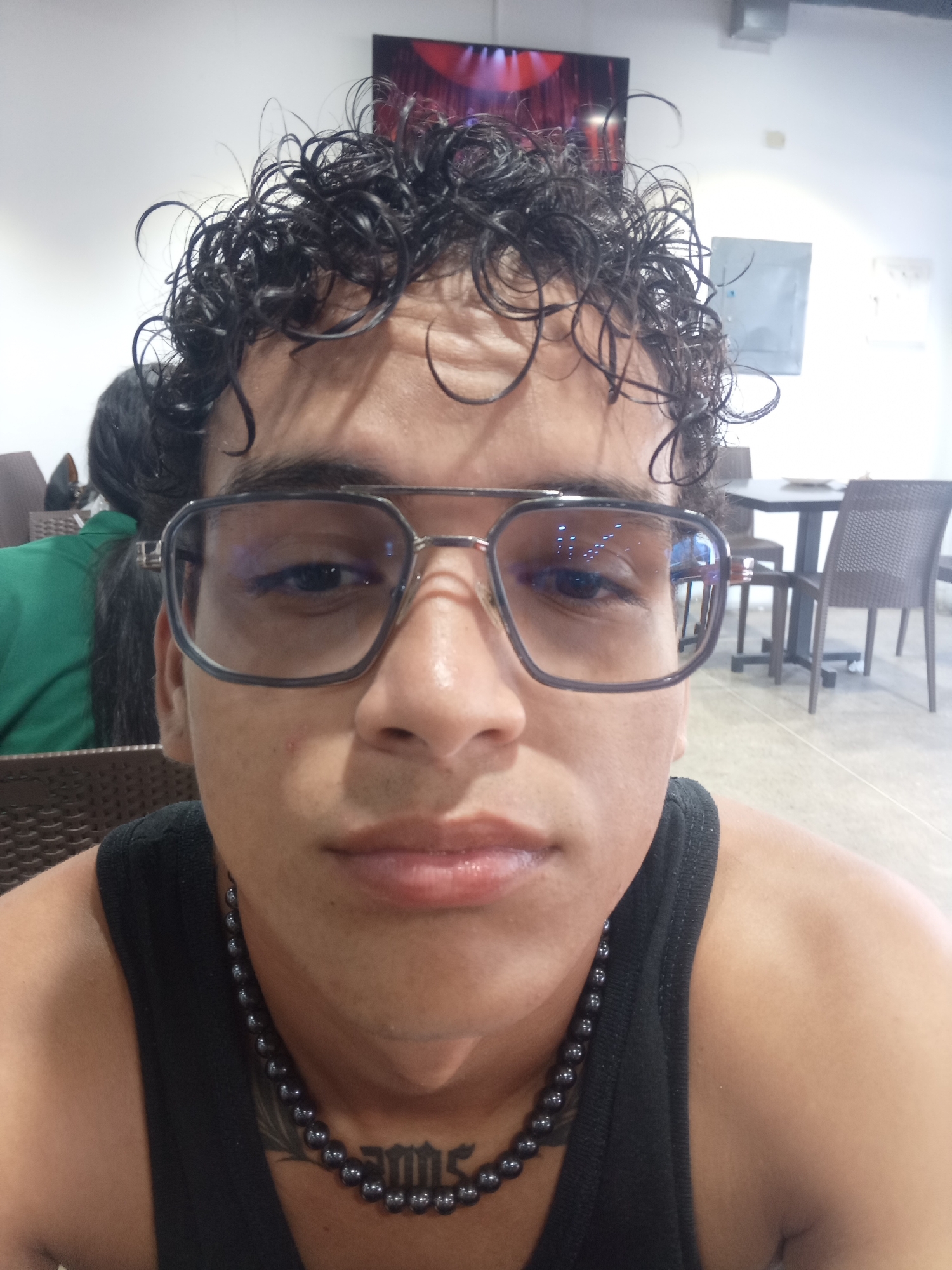 A close-up, selfie-style photograph shows a person with curly, dark hair and glasses. The person is wearing a black tank top with a beaded necklace and a tattoo on their chest. The background appears to be an indoor setting with tables and chairs, possibly a cafe or restaurant. The lighting is somewhat dim, with reflections visible in the glasses. The overall impression is a casual, candid shot. The location is identified as Juan Griego, Venezuela.