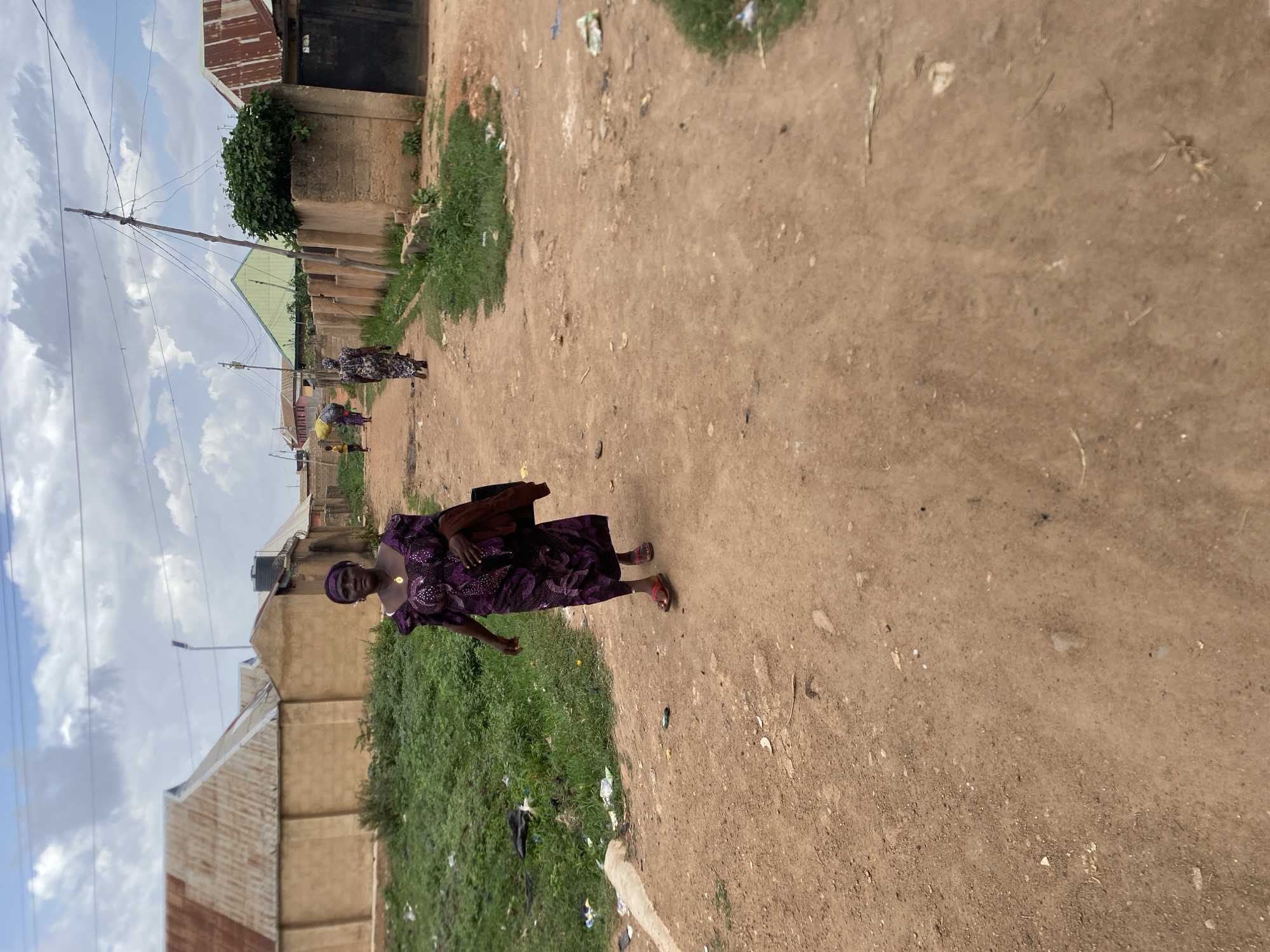 The image depicts an unpaved, dirt road in Bukuru, Nigeria. The road is bordered by buildings of modest construction and patches of grass containing scattered trash. A woman in a purple dress and headscarf walks along the road, carrying a brown purse or bag. Other individuals are visible further down the road. The sky is overcast with clouds.