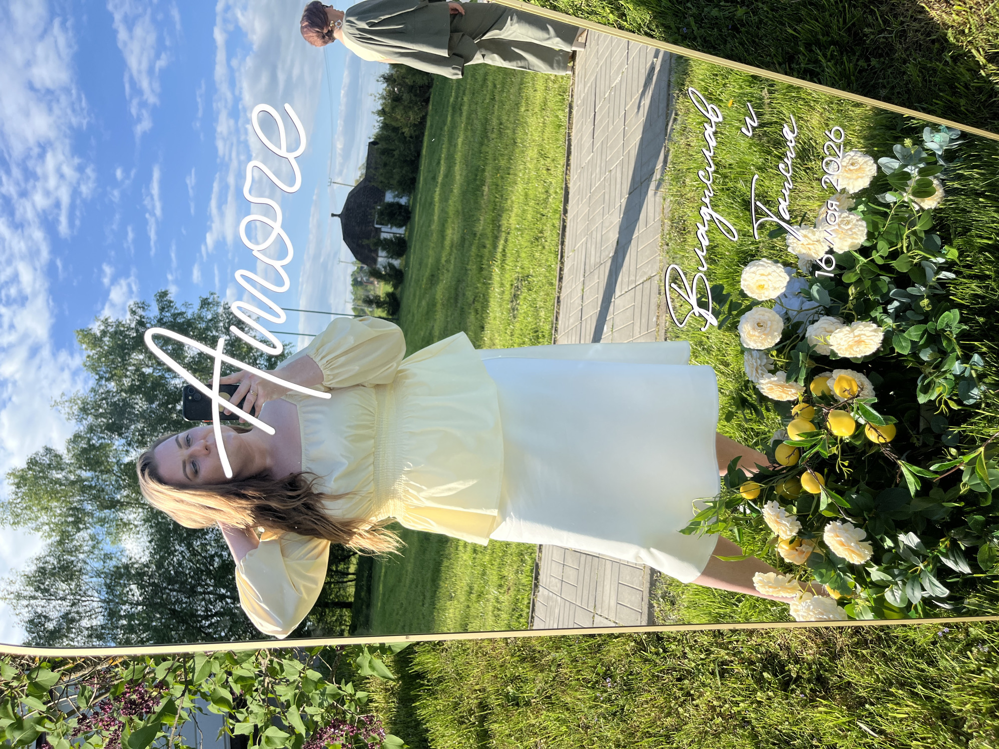A woman takes a selfie in a mirror outdoors on a sunny day. The mirror has gold trim and is decorated with flowers and greenery. In the mirror are the words "Amore," "Відчував," and "16.09.2026." A second person is reflected in the mirror in the background. The environment is grassy and appears to be a field or lawn near trees.