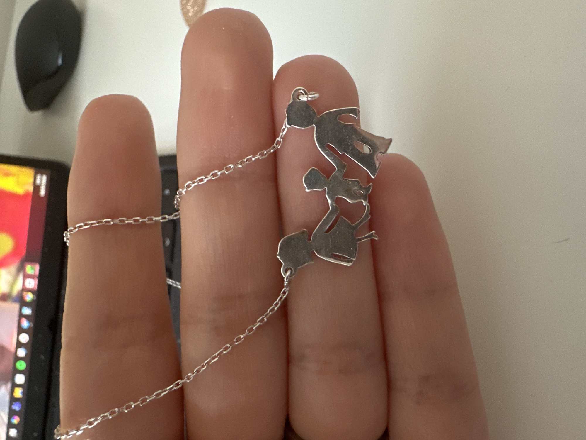 A silver necklace with a pendant depicting three stylized figures holding hands is being held between fingers against a blurred background. The figures represent a larger person on the left, a smaller person in the middle, and another larger person on the right. The pendant is suspended from a thin silver chain. The environment appears to be an indoor setting, possibly a desk area, with a computer monitor and a black mouse visible in the background. The lighting is natural. There is no information about the location in Leeds, United Kingdom.