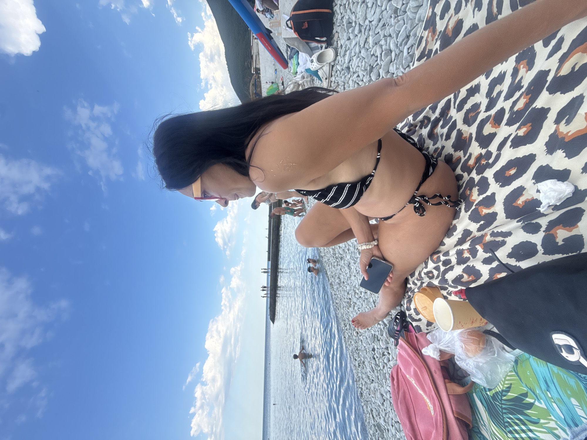A woman with dark hair, wearing a black and white striped bikini and yellow-tinted sunglasses, sits on a pebble beach in Partenit, Ukraine. She is holding a smartphone in her right hand. In front of her, on a leopard-print towel, are a pink bag, a black bag, a cup, and a plastic-wrapped item. The sea is to her left, with several people swimming and a pier in the distance. The sky is blue with scattered clouds.