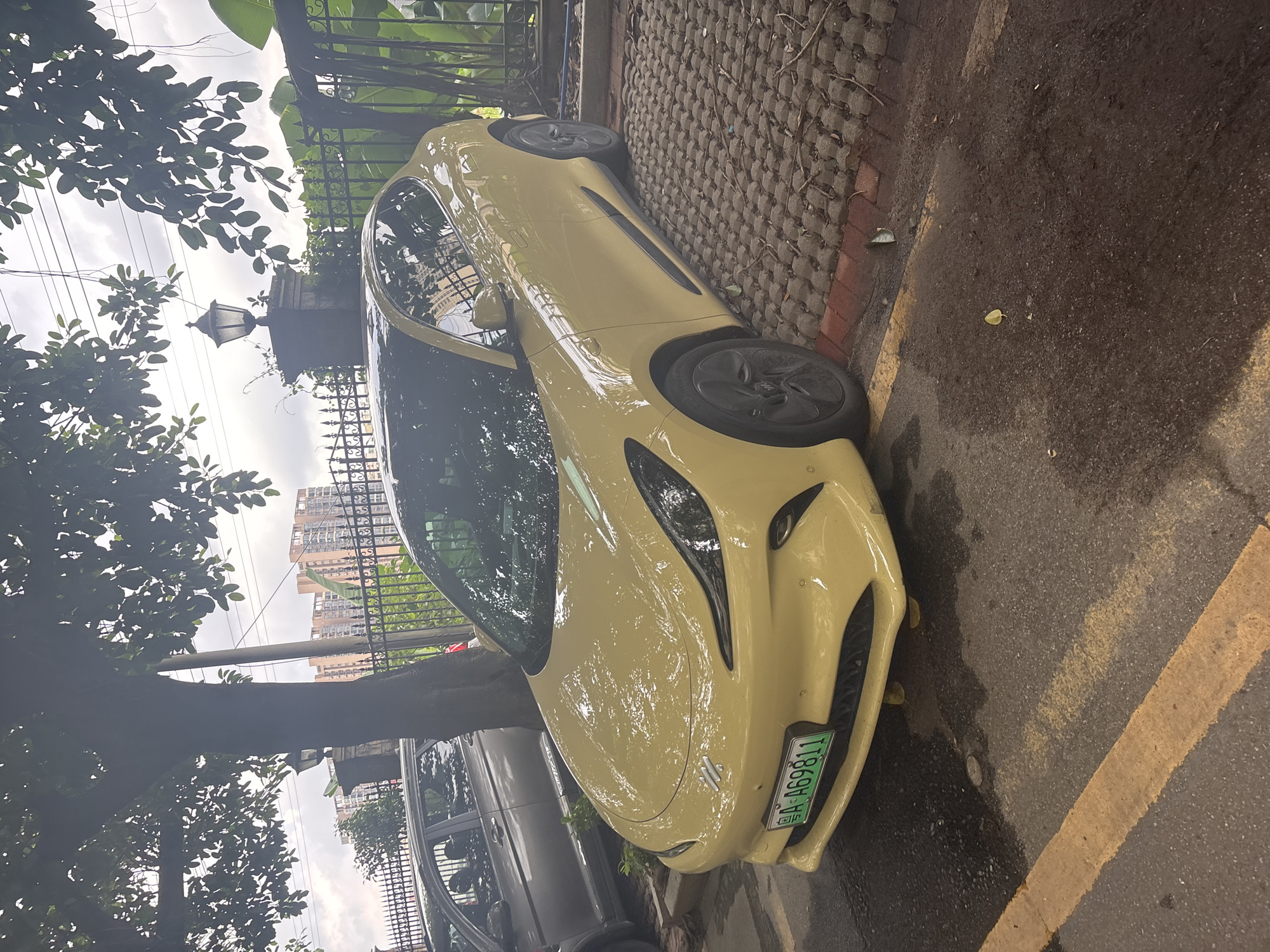 A yellow electric car is parked in an outdoor lot. The car is facing the right and its front license plate reads "A A69811". The car is parked on a surface of interlocking concrete pavers. Behind the car is a black metal fence and some lush green foliage. In the background, there are tall apartment buildings and a cloudy sky. To the left of the yellow car, a portion of a gray car is visible. The scene appears to be located in the Nanhai District of China.
