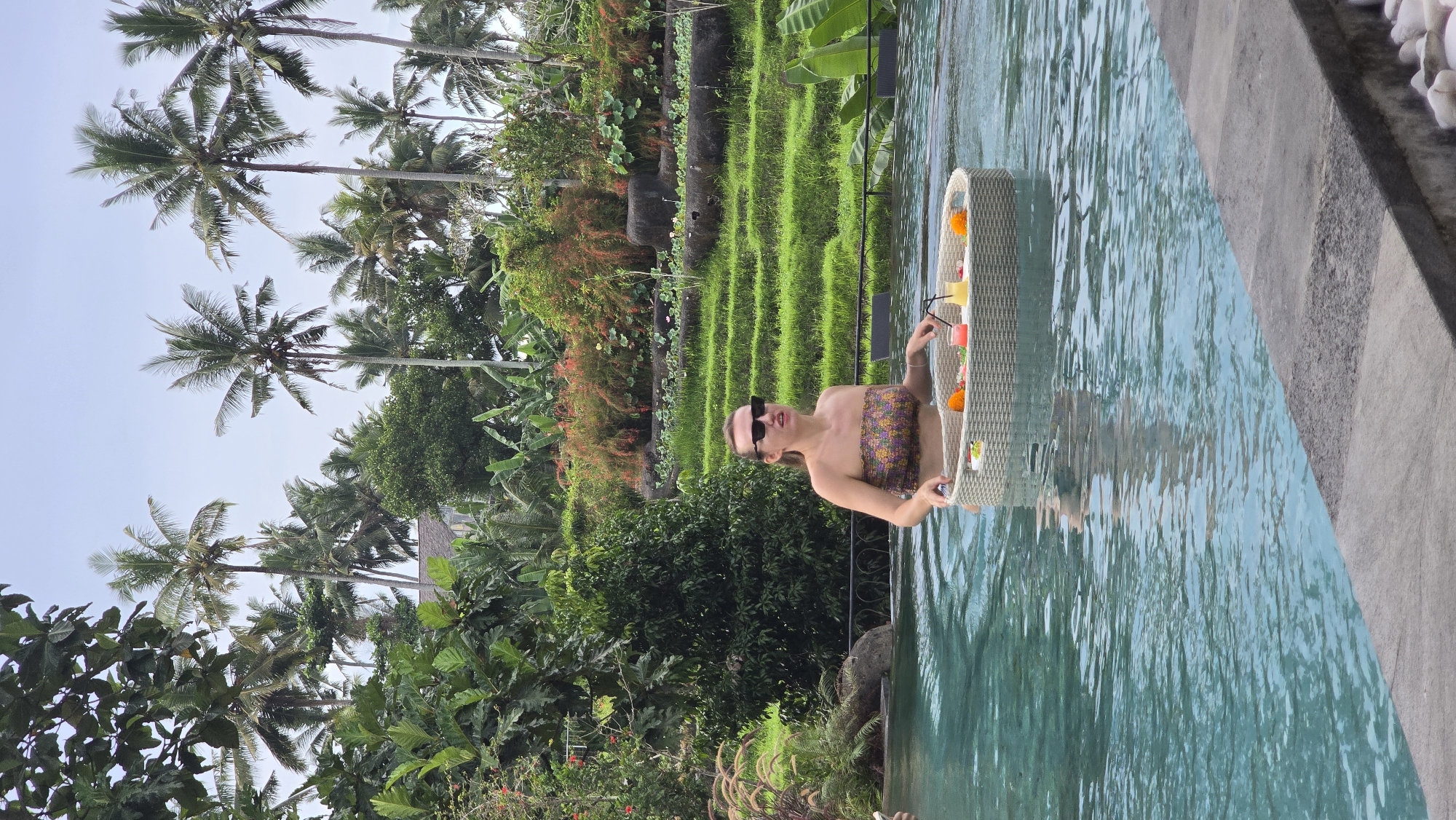A woman is captured relaxing in a swimming pool, enjoying a floating tray of drinks in a lush tropical setting, consistent with Ubud District, Indonesia. She wears a strapless, patterned top and dark sunglasses, with her hair neatly pulled back. Her hands are near the white wicker-style floating tray, which holds several vibrant, colorful drinks with straws and garnishes, along with some orange decorative elements.
The pool water is clear and inviting, reflecting the bright daylight. To the right, a gray concrete pool edge with small white pebbles adds a clean border to the scene. The background is dominated by a rich, verdant landscape. Tall palm trees, characteristic of tropical regions, ascend towards a bright, clear sky. Below them, dense layers of green foliage, bushes, and trees create a natural, vibrant environment. Further back, distinctive terraced rice paddies, filled with bright green crops, are clearly visible, serving as a strong geographical cue for Indonesia. Some of the distant vegetation shows hints of reddish-brown hues. The scene depicts a serene and leisurely moment, likely at a resort, under pleasant, sunny daytime weather. There is no visible text in the image.