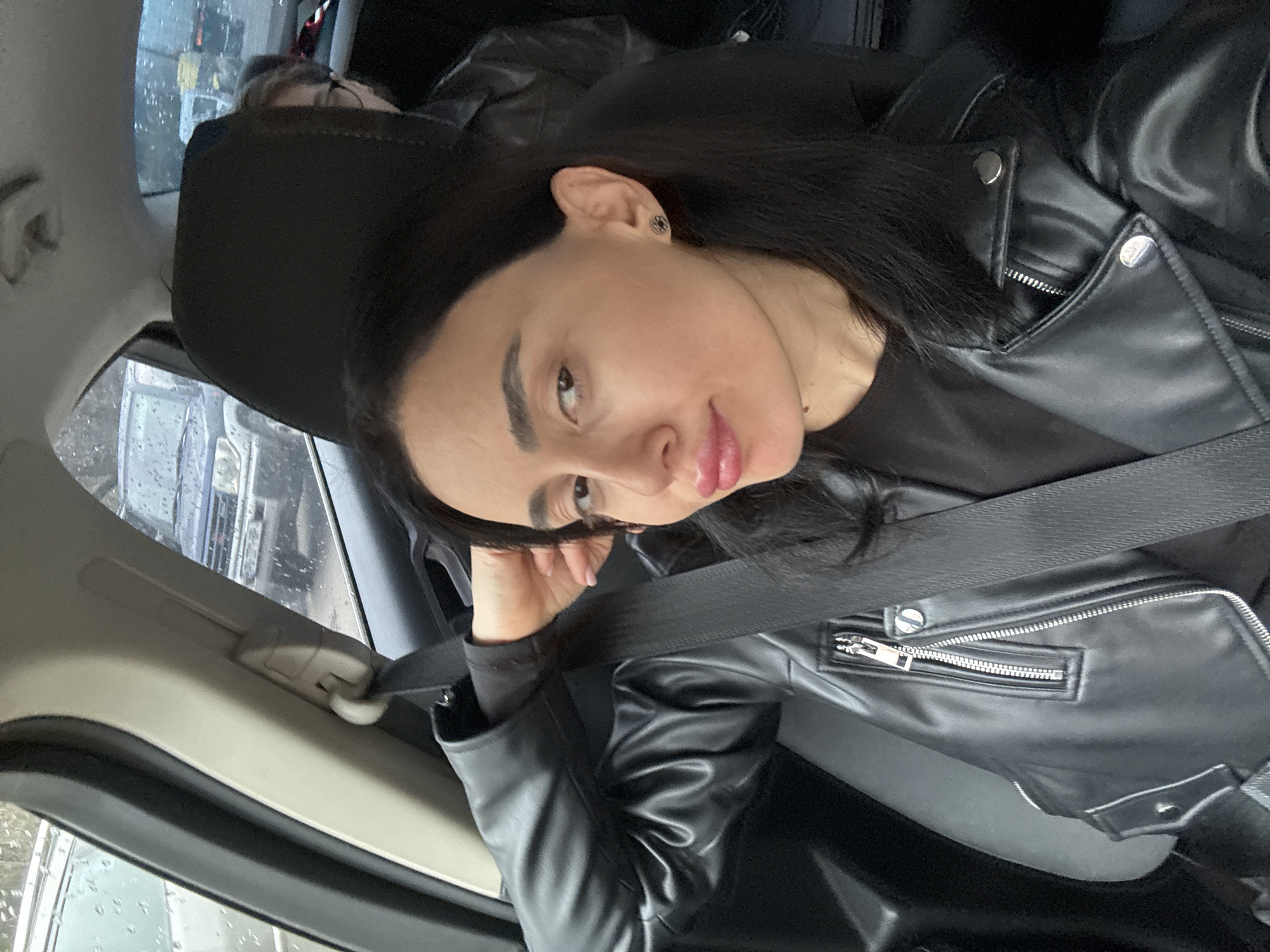 A woman with long dark hair, wearing a black leather jacket and a black shirt, is sitting in a car. She is wearing a seatbelt. Her left hand is near her face. The background shows a blurry view of a street with other cars and wet conditions, suggesting rain. Given the setting and the appearance of the woman, the location might be Tbilisi, Georgia, but this cannot be definitively confirmed from the image.