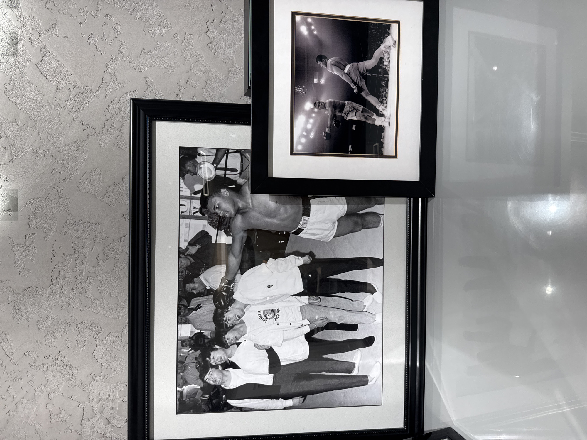 The image displays two black and white framed photographs, mounted on a light gray wall with a distinctive textured finish, possibly stucco or Venetian plaster. The entire scene is captured with the camera rotated 90 degrees clockwise, making the horizontally oriented photographs appear vertical in the provided image.
The smaller, upper frame contains a dynamic black and white photograph of a boxing match in progress. Two boxers are shown inside the ring, illuminated by bright overhead lights. One boxer, in light-colored shorts, is leaning forward, suggesting an active moment in the fight. The opponent, in darker shorts, is also visible. Spectators can be faintly seen in the background, adding to the atmosphere of an intense sporting event.
The larger frame, positioned below the smaller one, showcases another black and white photograph featuring legendary boxer Muhammad Ali. He is depicted shirtless, wearing white boxing shorts with the brand "EVERLAST" clearly visible on the waistband. Ali appears to be interacting with a diverse group of people surrounding him, possibly in a locker room, during a public appearance, or a training session. Many onlookers are gazing at him with expressions of admiration and excitement. One person close to Ali is holding a cup. Among the crowd, a person wearing a white long-sleeved shirt has text visible on it, reading "SUPREMA" and "RACE WRIST". The composition suggests a moment of close engagement between Ali and his admirers.
The photographs are presented with thick white mats inside sleek black frames. A reflective white surface is visible to the right of the frames, exhibiting some glare. This display of iconic boxing imagery is situated in Dubai, United Arab Emirates.