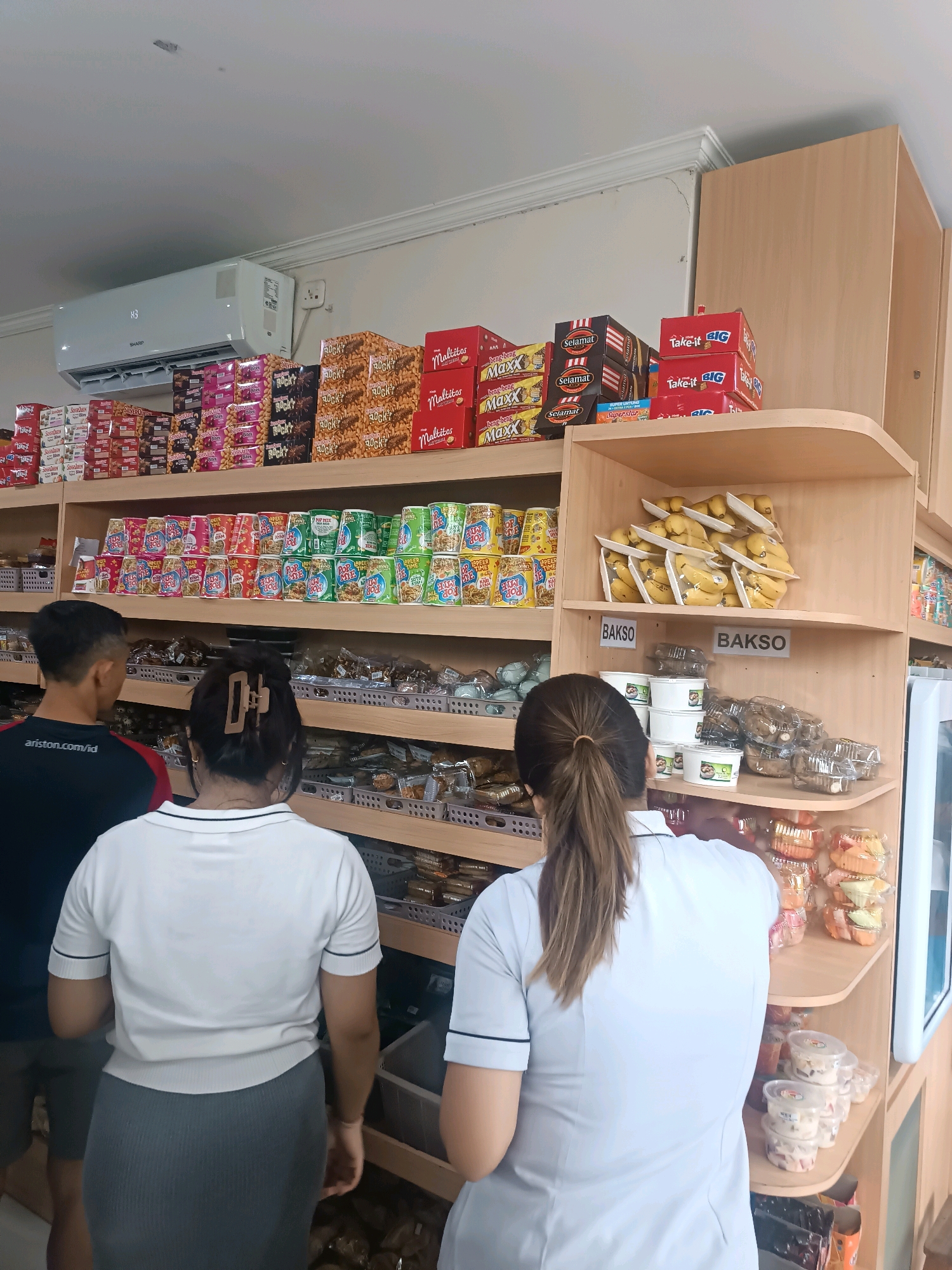The image depicts the interior of a small convenience store, likely a warung or minimarket, in Denpasar, Indonesia. The store is well-stocked with various consumer goods, including packaged snacks, drinks, and some fresh produce. In the foreground, three individuals are visible. Two women, one with her back to the camera and wearing a white polo shirt and grey skirt, and another wearing a light blue polo shirt, are browsing the shelves. A man, wearing a dark t-shirt with "ariston.com/id" visible on the back, stands to the left, also looking at the products.
The shelves are made of light-colored wood. The upper shelves are filled with numerous boxes of snacks and sweets, including "Maltitos," "Maxx," and "Selamat" branded items. Some boxes are stacked neatly, while others are more loosely arranged. On the right side of the upper shelves, packages of bananas are displayed. Below these, there are shelves displaying more packaged goods, including what appear to be instant noodles or similar dry food items in colorful packaging. Further down, shelves hold clear plastic containers filled with various food items, some of which are labeled "Bakso," suggesting meatballs or related products. The store also features an air conditioning unit mounted high on the wall above the shelves. The overall environment suggests a typical Indonesian local shop.