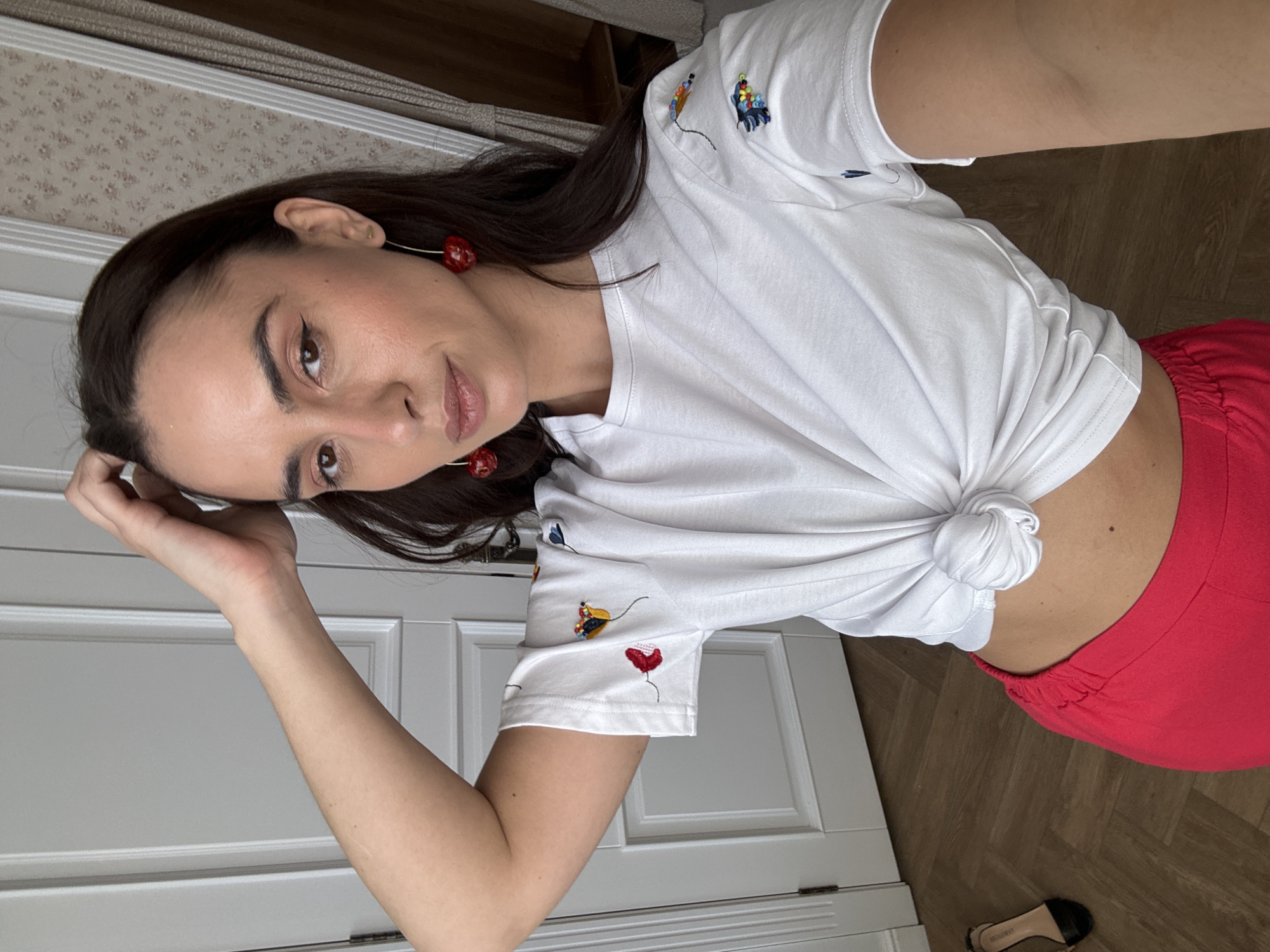 An indoor selfie captures a young woman, likely in her 20s or 30s, in what appears to be a residence in Krasnoyarsk, Russia. The image is rotated 90 degrees clockwise, showing the woman positioned diagonally across the frame, looking directly at the camera with a neutral expression. She has long, dark brown hair and is wearing natural-looking makeup, including winged eyeliner and nude lipstick.
Her attire consists of a white short-sleeved t-shirt with small, colorful embroidered designs on the sleeves and shoulder, possibly depicting flowers or abstract shapes. The t-shirt is tied in a knot at her waist, exposing a sliver of her midriff. She is also wearing bright red pants or shorts with ruched elastic detailing at the waist. Large, red cherry-shaped dangle earrings with gold-toned wires accessorize her look. Her left hand rests near her head, suggesting she is posing. A small mole is visible on her lower back/side.
The background reveals a home environment. To her left, a white paneled door or cabinet is visible. The floor is made of dark wood with a herringbone pattern. In the upper left corner, there's a light-colored wallpaper with a subtle pattern. A black flat shoe with a light-colored sole, displaying the text "MSGM", is seen on the wooden floor. The soft lighting suggests it is daytime.